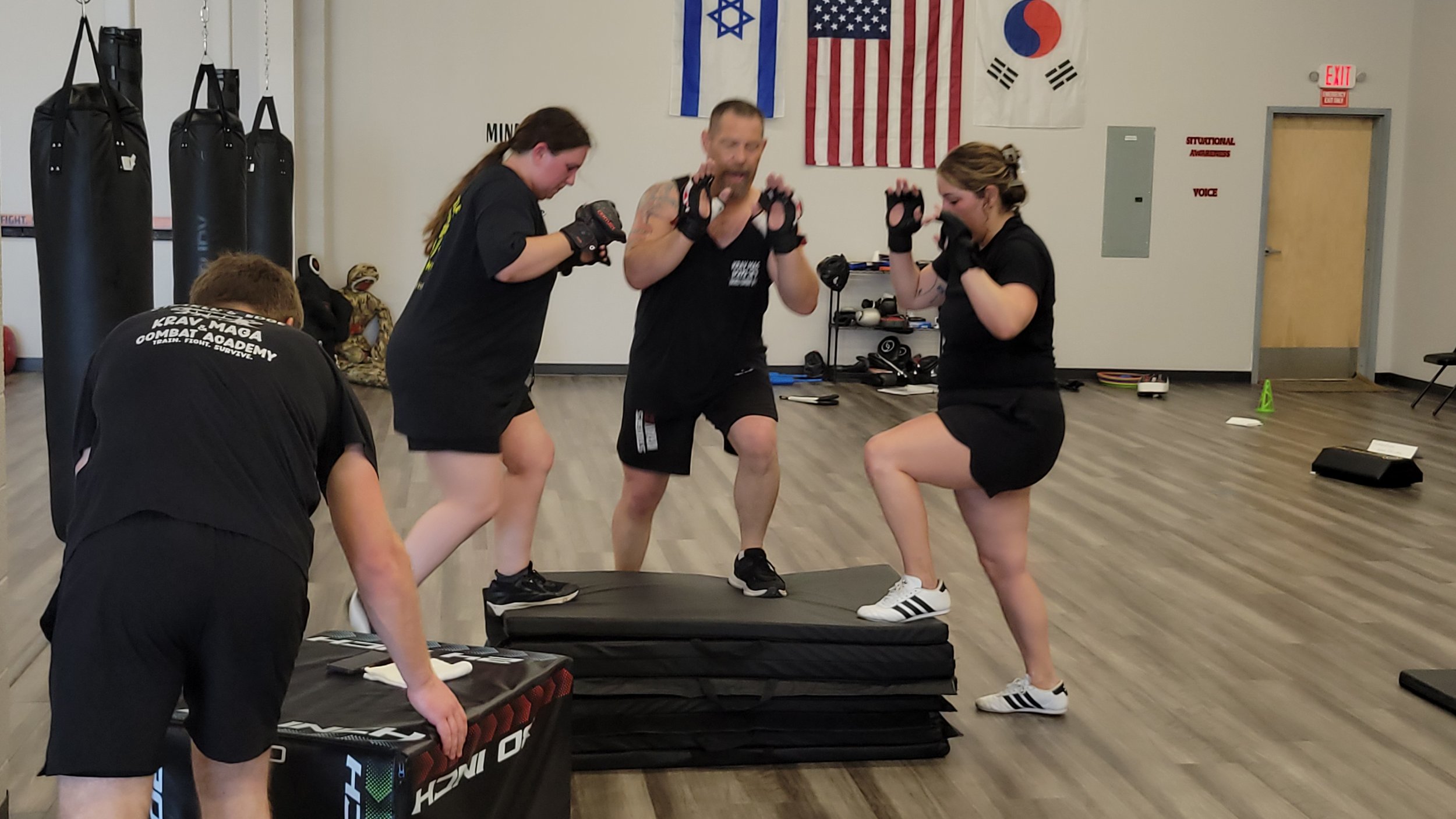 Four people in workout attire are training in a martial arts gym with boxing bags, flags on the wall, and fitness equipment in the background. Two women are stepping onto padded platforms while a man appears to be coaching them.