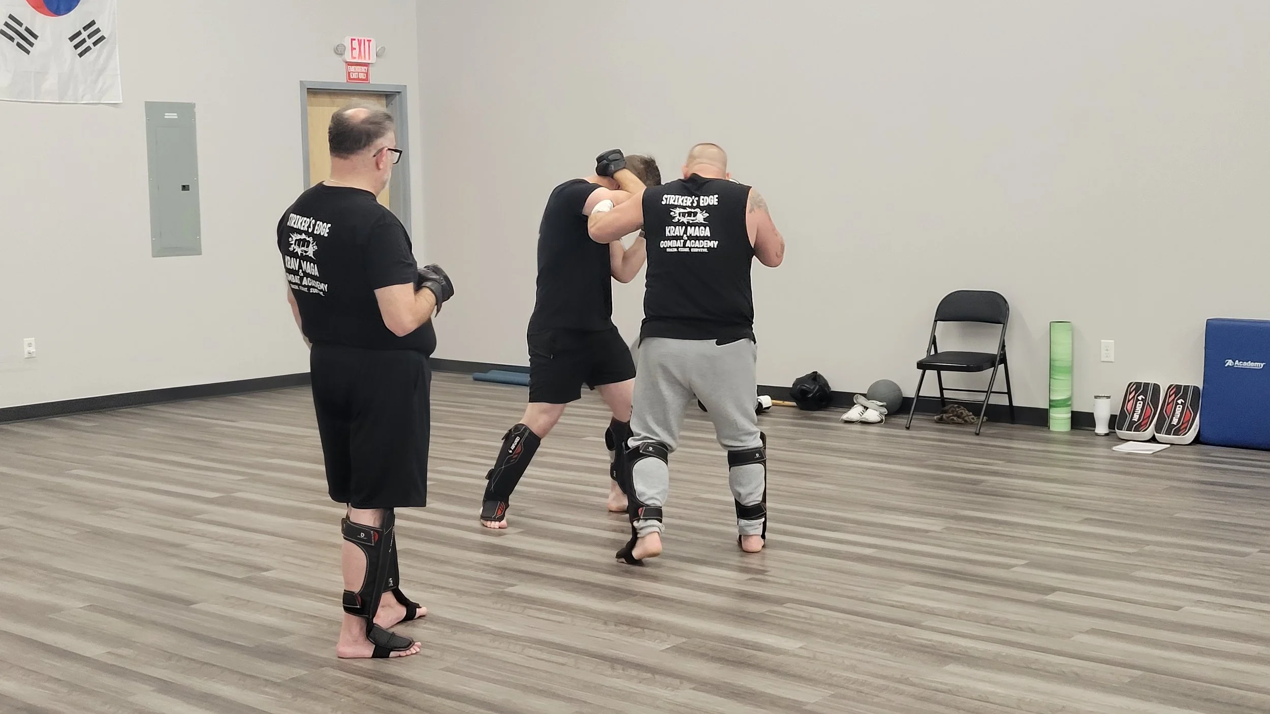 Three men in a martial arts classroom practicing sparring while wearing protective gear, with one person coaching or observing.