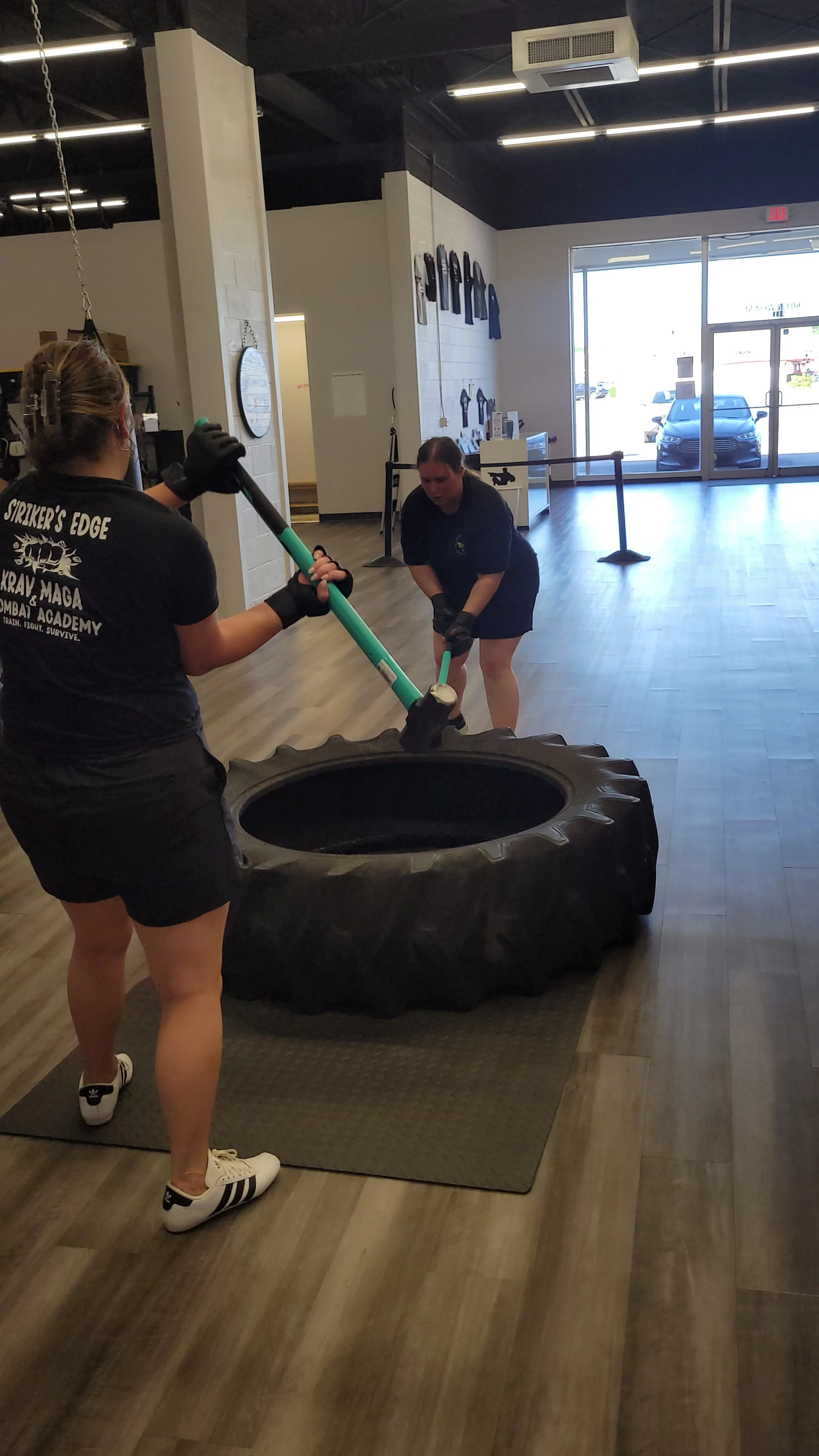 Two women in a gym-like setting are preparing to jump into a large tire for training. One woman is holding a green pole and is wearing a black t-shirt with text on the back, black shorts, and white sneakers with three black stripes. The other woman is bending over, holding a green pole, dressed in black shorts, and a black T-shirt.