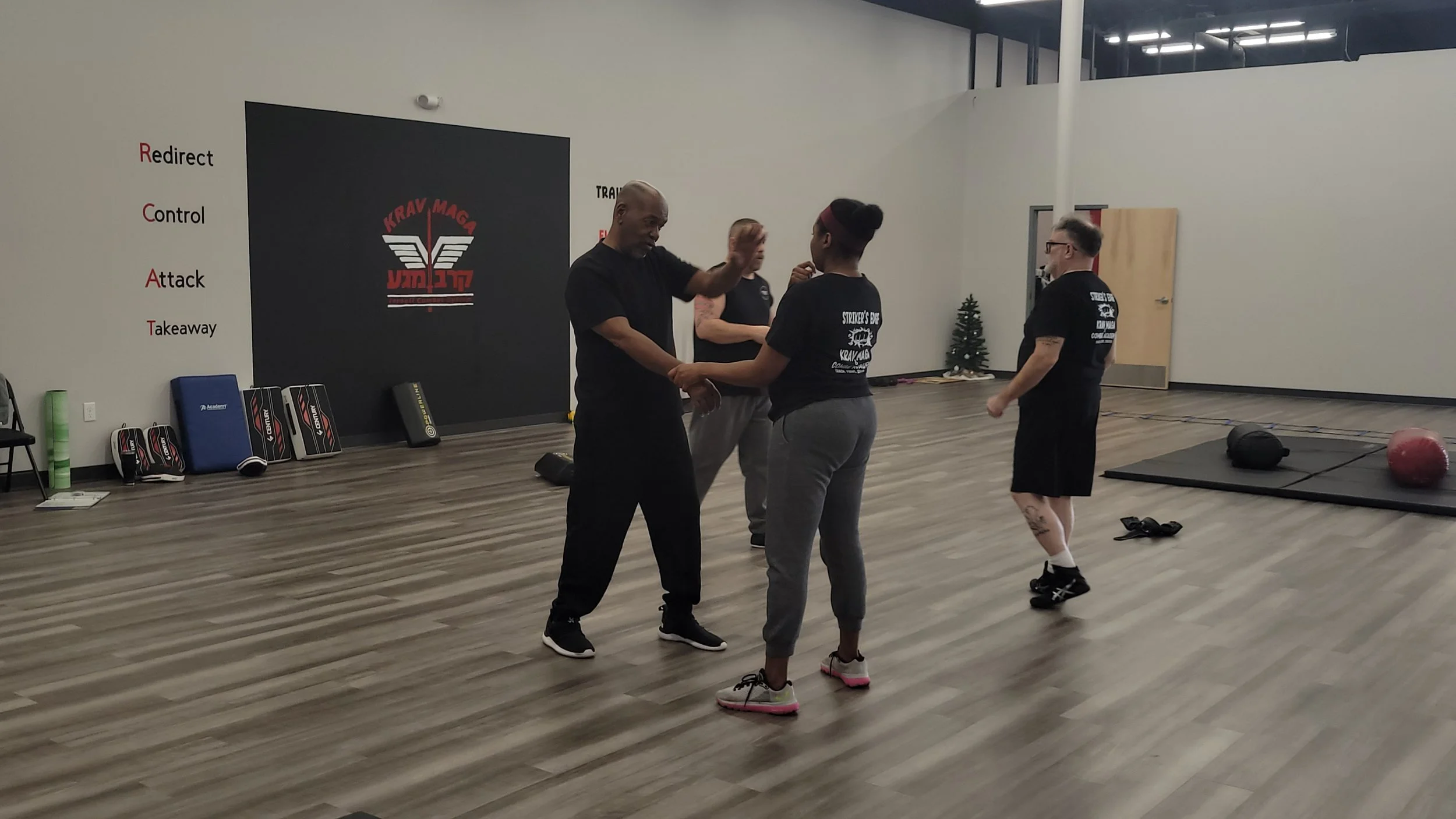 Martial arts instructor practicing techniques with a woman in a martial arts training room, with other participants in the background and training equipment along the walls.