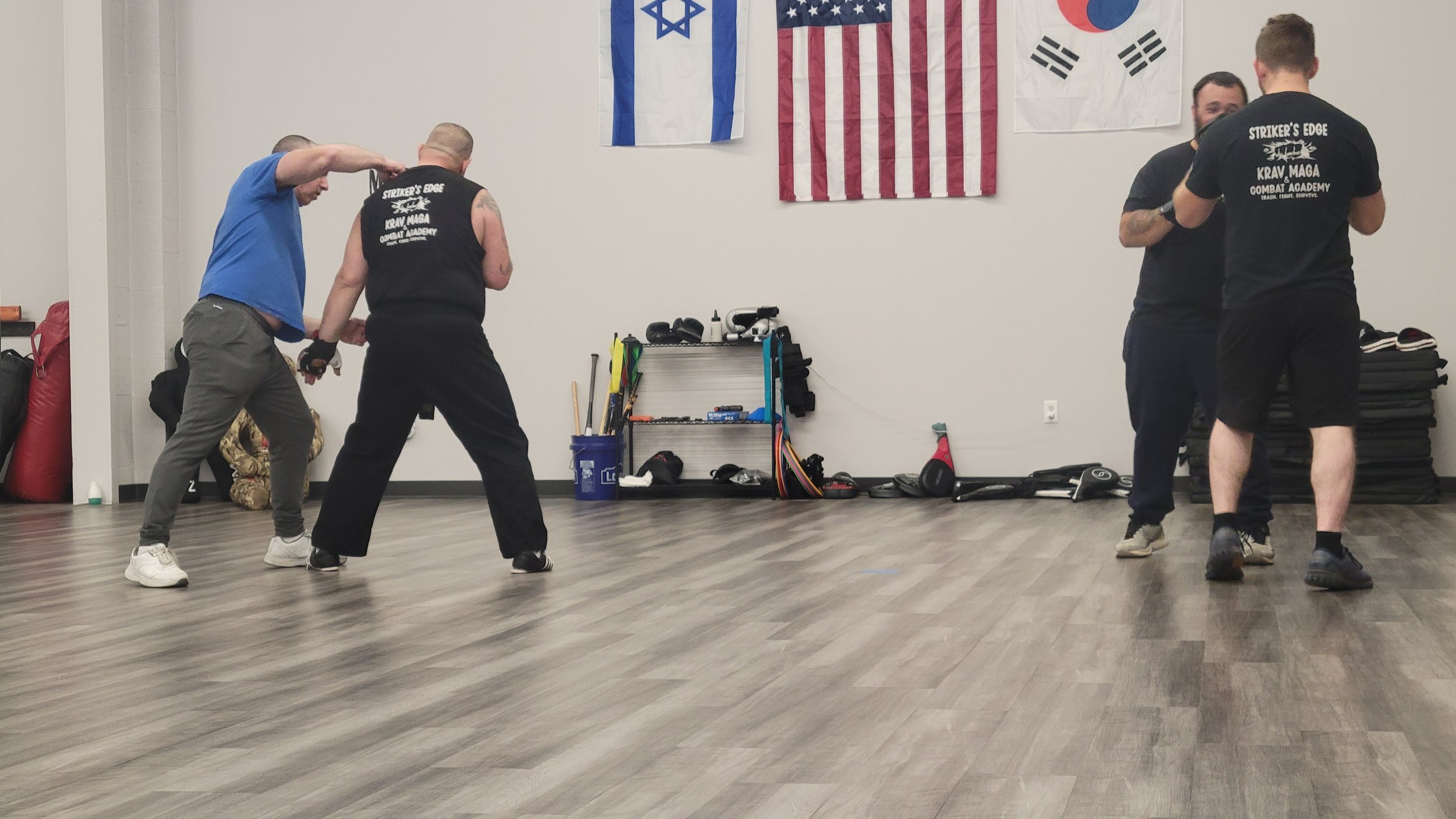 Four men practicing self-defense or martial arts in a gym with American, Israeli, and South Korean flags hanging on the wall.