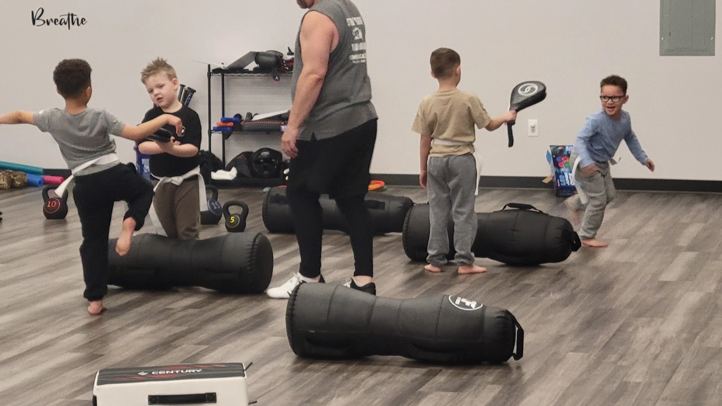 Kids and a woman in a martial arts gym, engaging in martial arts training with punching bags and foam rollers on the floor.