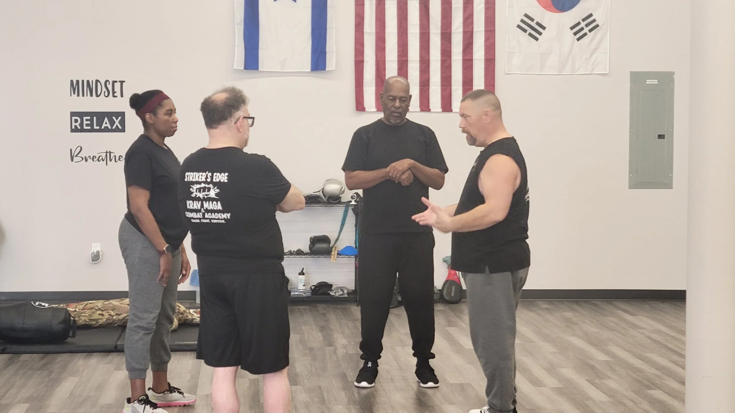 Four people in a martial arts training studio having a discussion. Two men and one woman are listening to a man explaining techniques. The woman and one man are wearing black t-shirts, and the other man is shirtless with a sleeveless black shirt. Behind them, there are three flags on the wall, including the American flag, the South Korean flag, and a flag with blue and white stripes. The wall has a motivational quote: 'Mindset Relax Breathe'. There is gym equipment and a placard with training gear on a shelf.