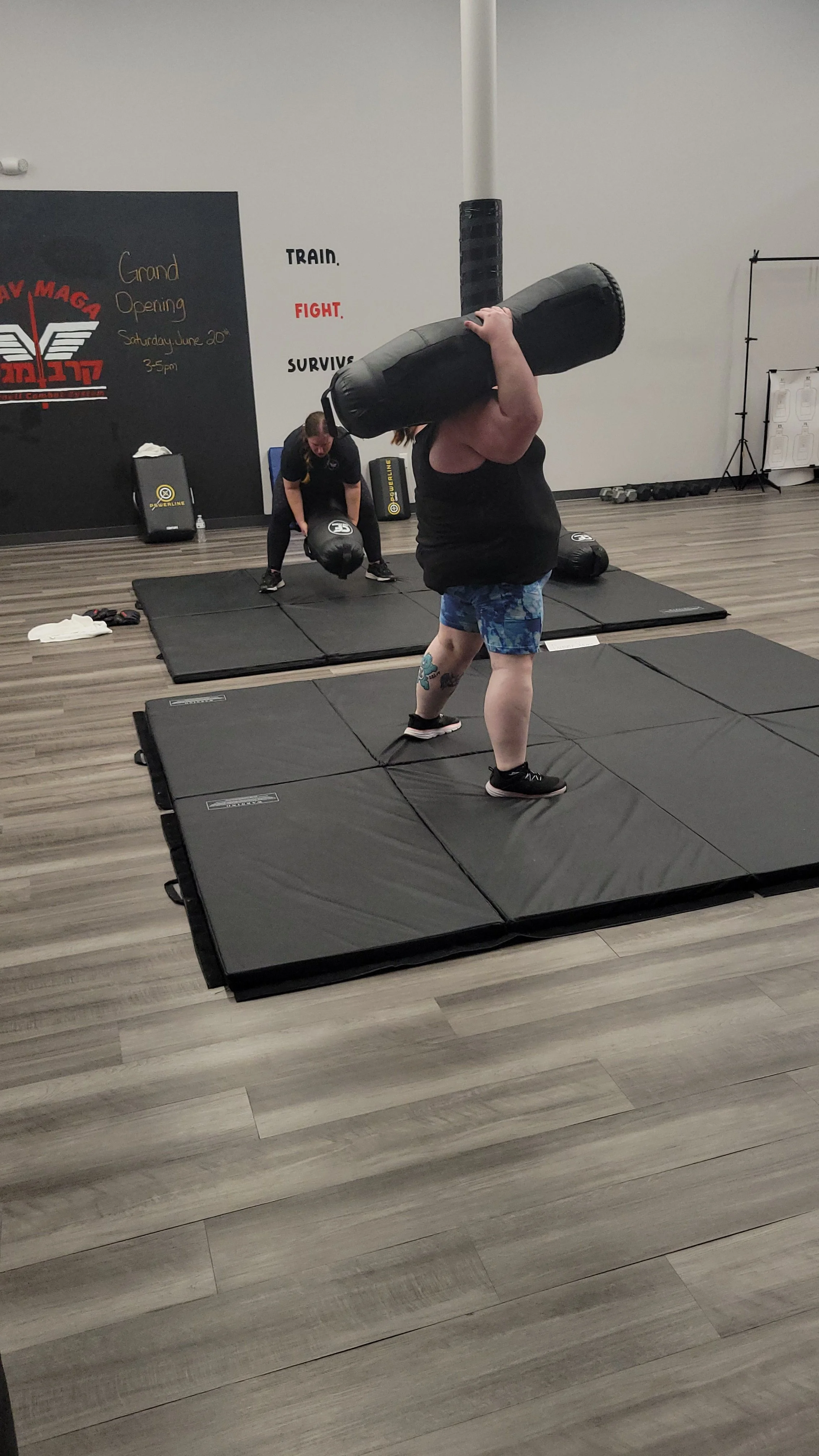 A person lifting a large black punching bag in a gym. There are padded mats on the floor and other gym equipment in the background.