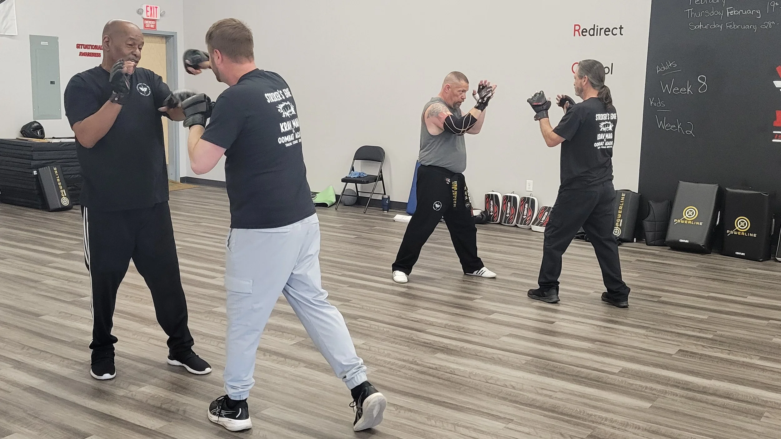 Four men practicing martial arts in a training room. Two men are sparring with gloves, while the other two are in stance, ready to train or spar. The room has wooden floors, a black wall with white and red text, and fitness equipment in the background.