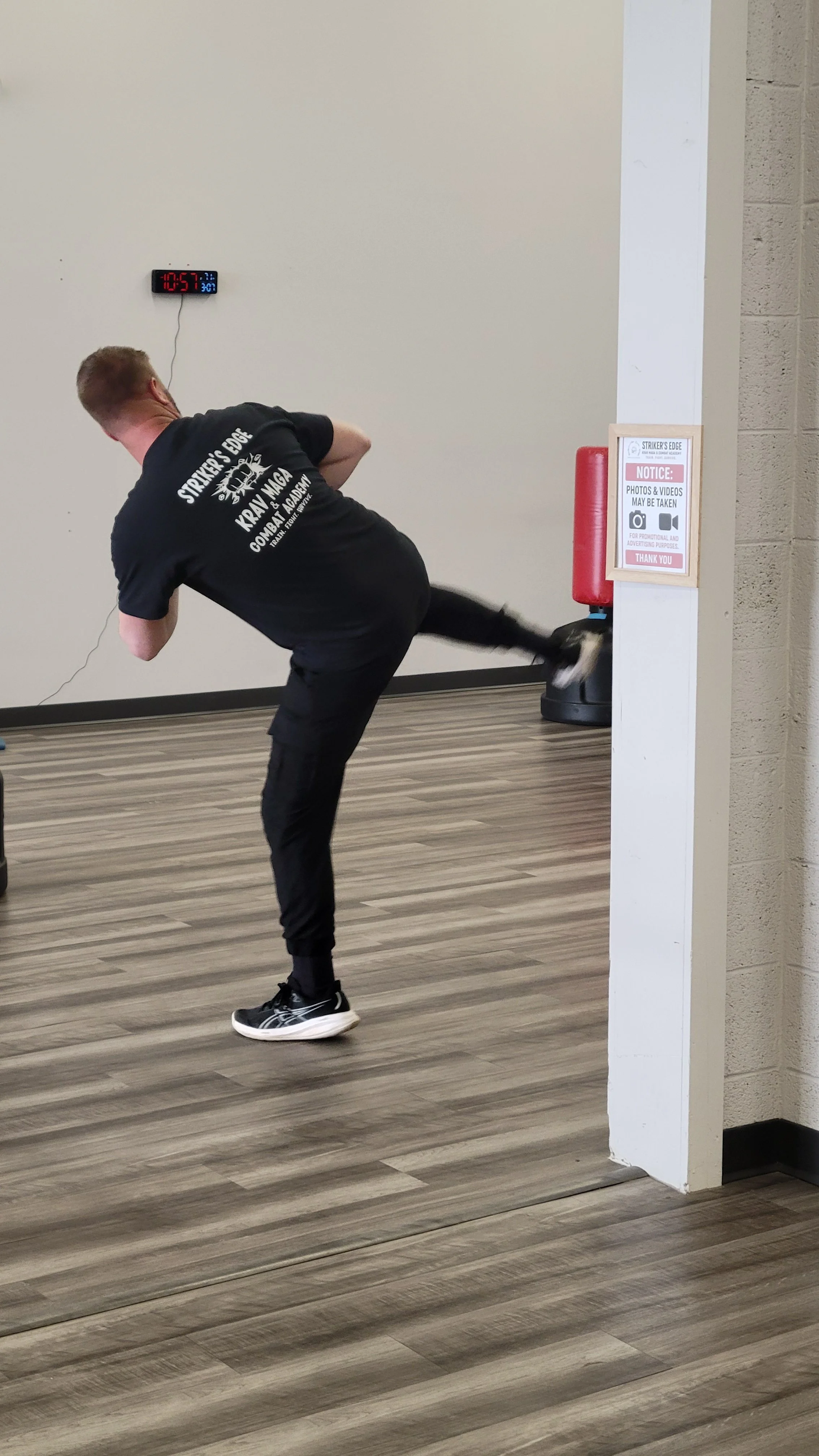 A man in black workout clothes is performing a high kick during a martial arts demonstration at a gym.