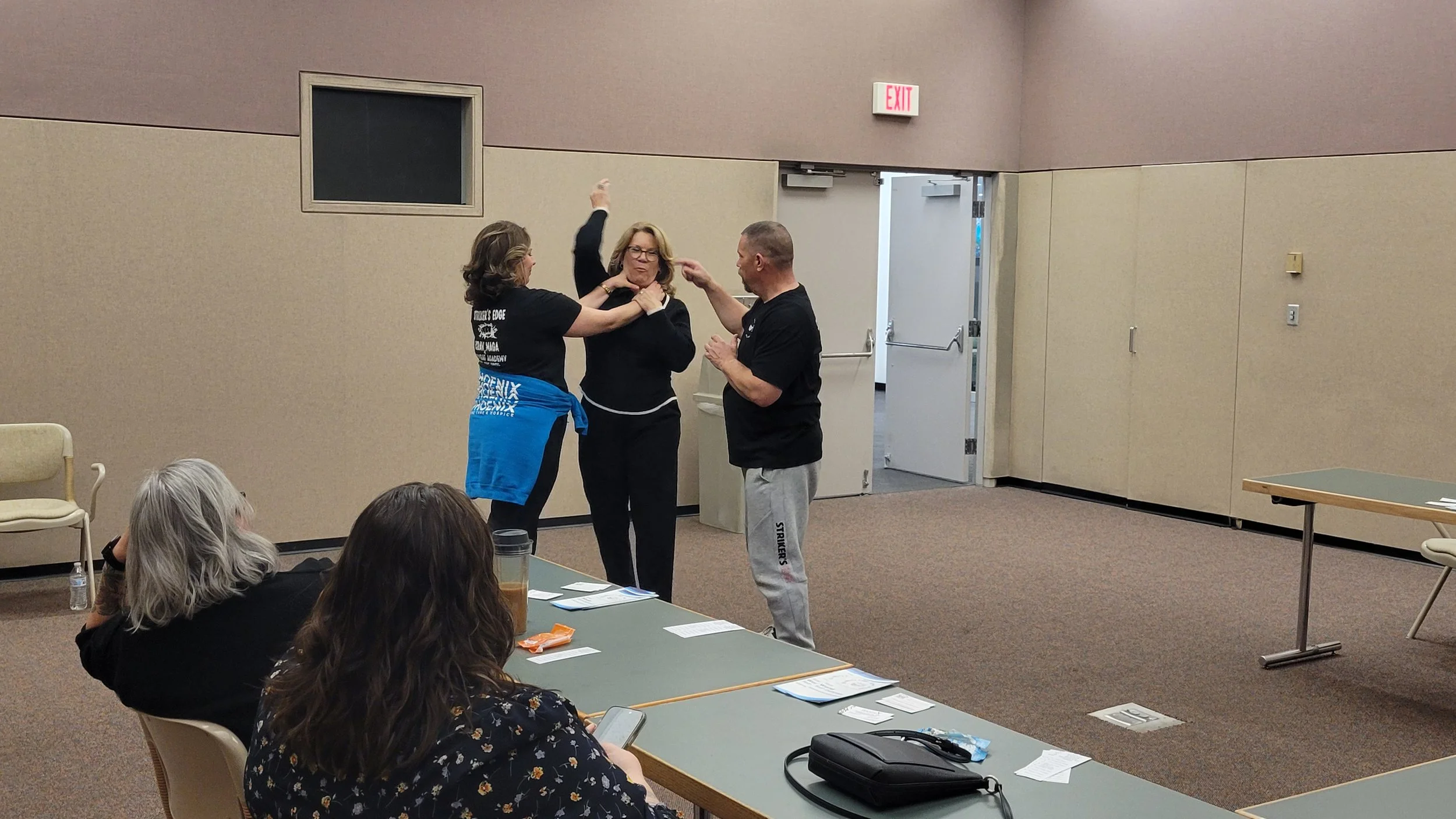 Three people fight in a room, two women and a man, while two seated women watch and use their phones. The room has beige walls, chairs, tables, and an open exit door.