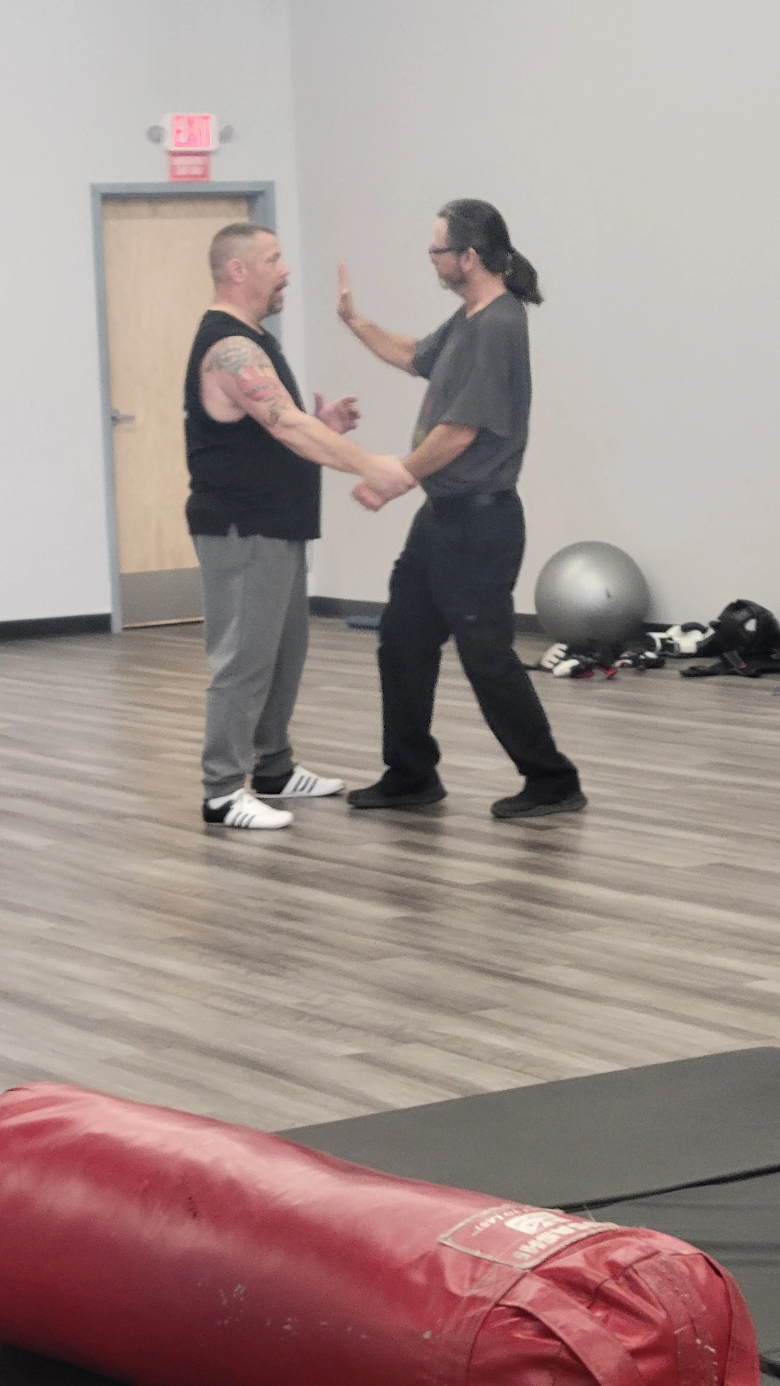 Two men are practicing a martial arts or self-defense move in a gym or training area. One man is holding the other's wrist while they face each other, with their hands touching. The man on the right is wearing glasses, a gray T-shirt, and black pants, while the man on the left has short hair, tattoos on his arm, and is wearing a black sleeveless shirt and gray sweatpants. Training gear like a medicine ball, gloves, and a punching bag are visible on the floor, with a door and an exit sign in the background.
