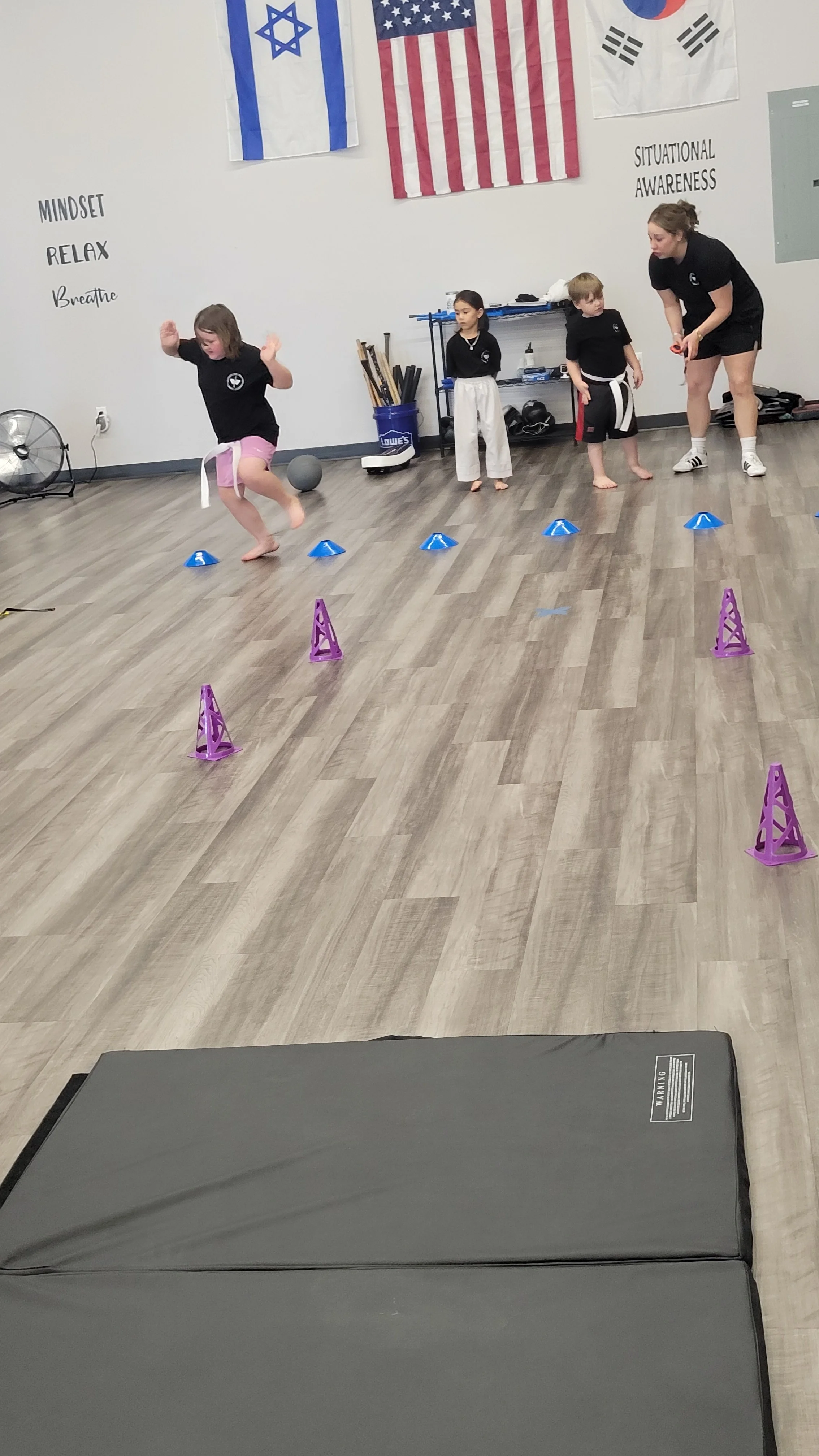 Children practicing martial arts in a dojo with American, Israeli, and South Korean flags hanging on the wall. One child is jumping over small blue and purple cones as an instructor supervises.