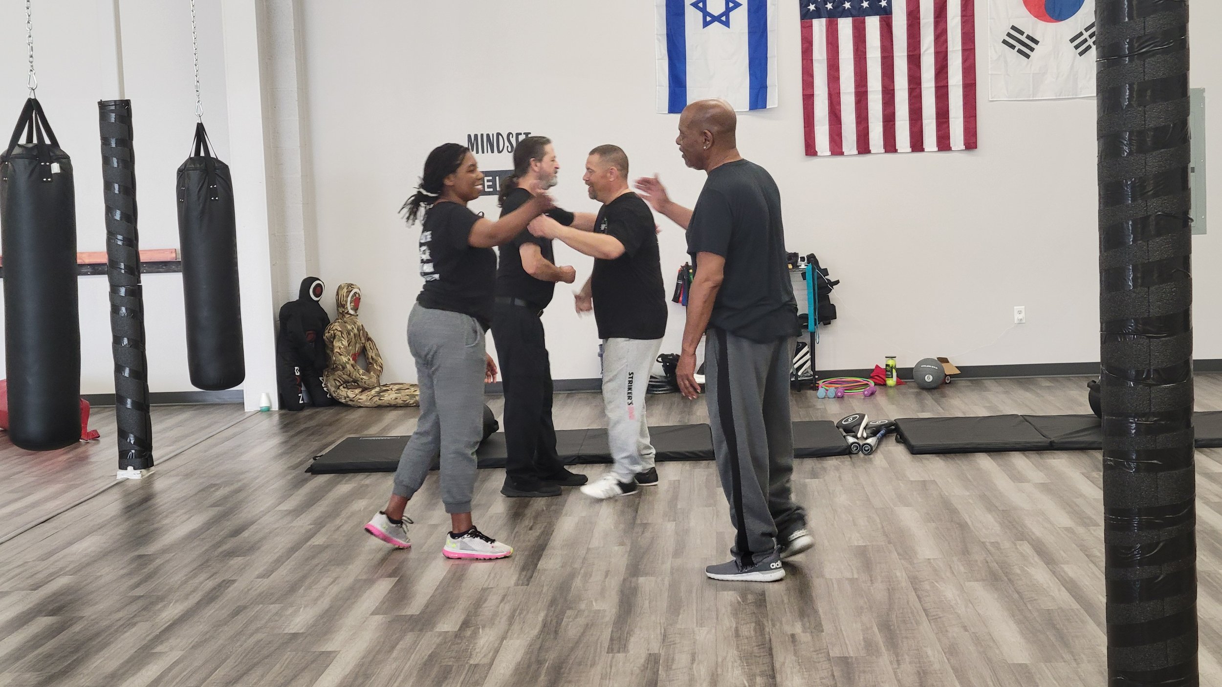 Five people in a gym engaging in martial arts training, with punching bags and flags on the wall.