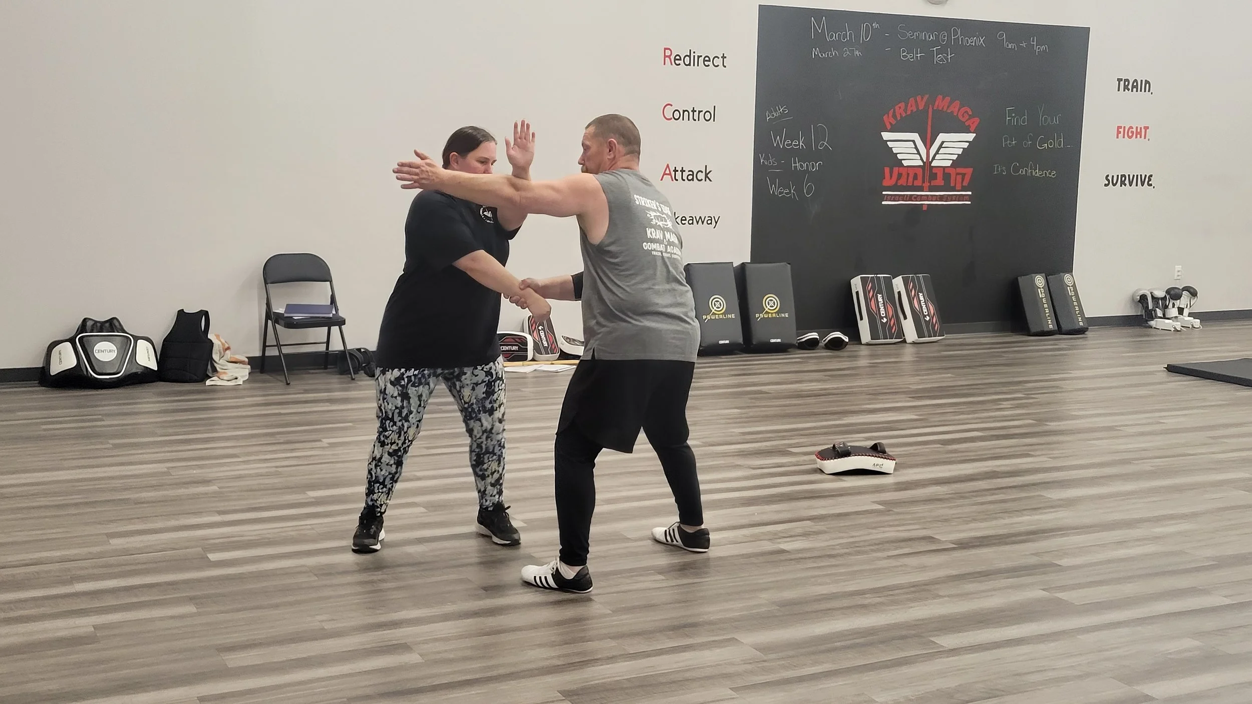 Two people practicing martial arts in a gym. The woman is wearing a black shirt and patterned leggings, and the man is wearing a gray sleeveless shirt and black shorts. They are engaged in a self-defense demonstration.