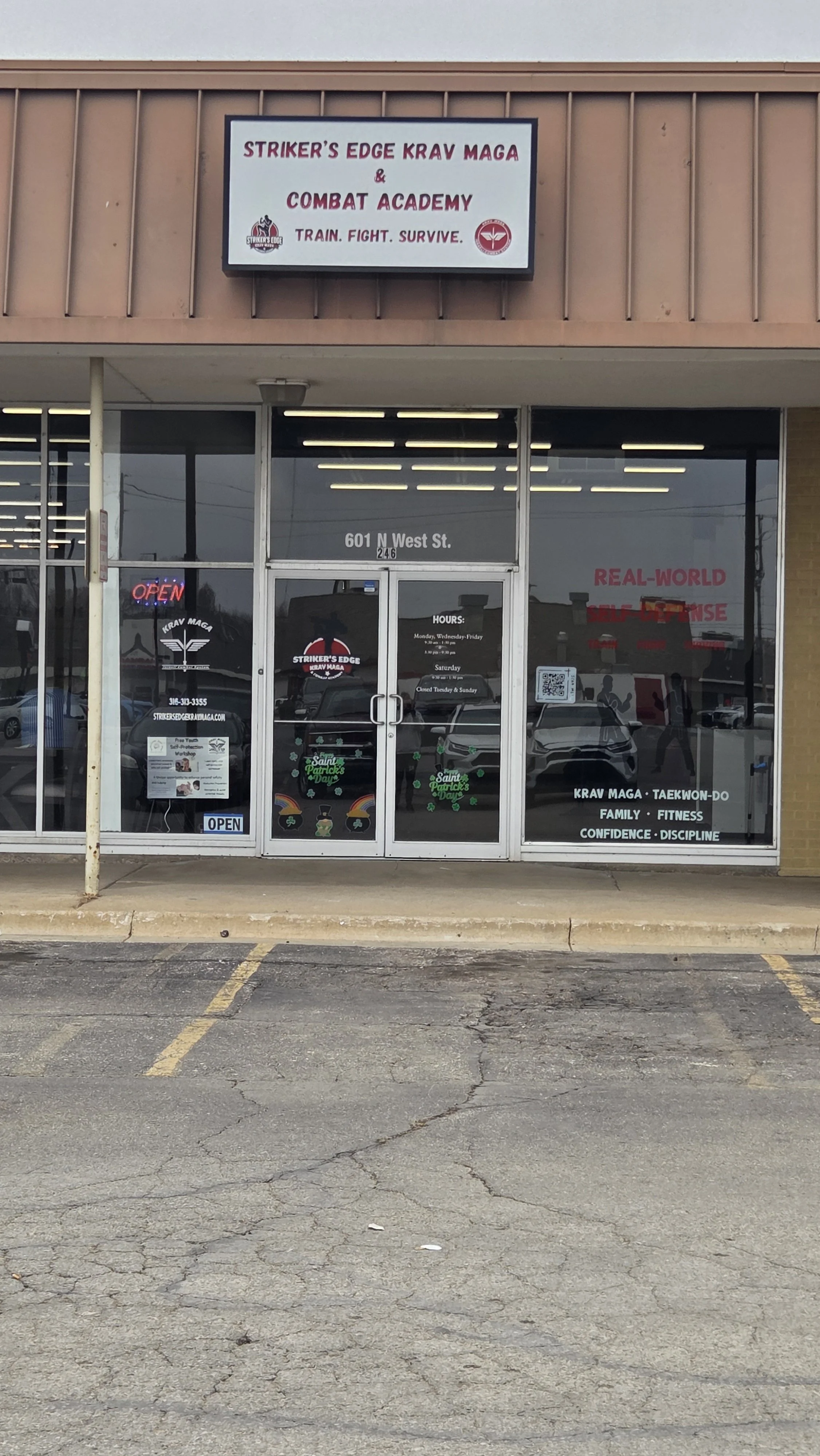 Front entrance of a martial arts gym with large glass windows and door, signage displaying the gym's name, operating hours, and services like Krav Maga, Taekwondo, family fitness, confidence, and discipline. The building has a signboard on top with t