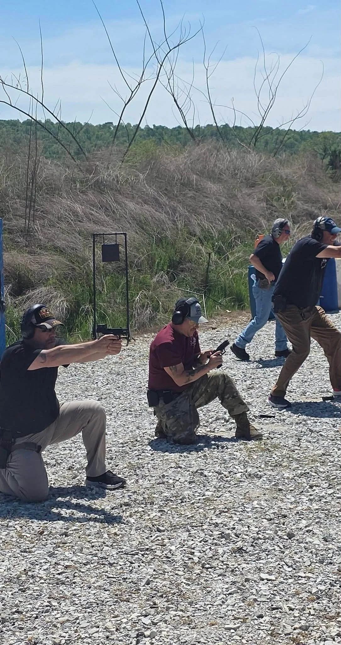 Five individuals at a shooting range, some kneeling and others standing, aiming firearms toward targets with ear protection and firearms handling safety.