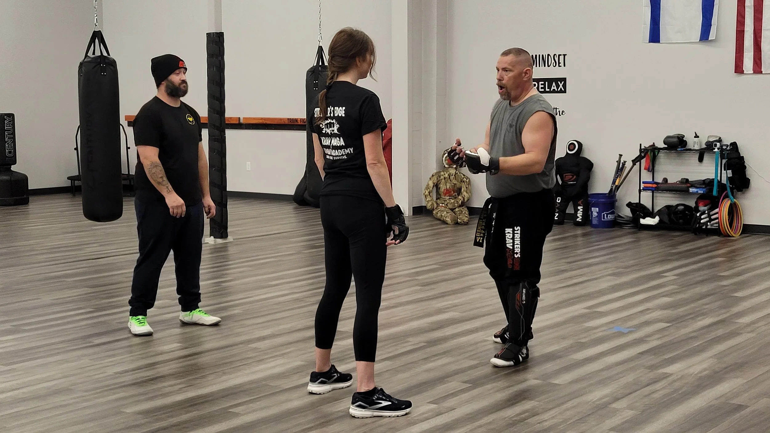 Three people in a martial arts gym, two men and a woman. The woman in the center is holding hand wraps, talking to a shirtless man wearing martial arts gear. The third man is standing behind them, observing.