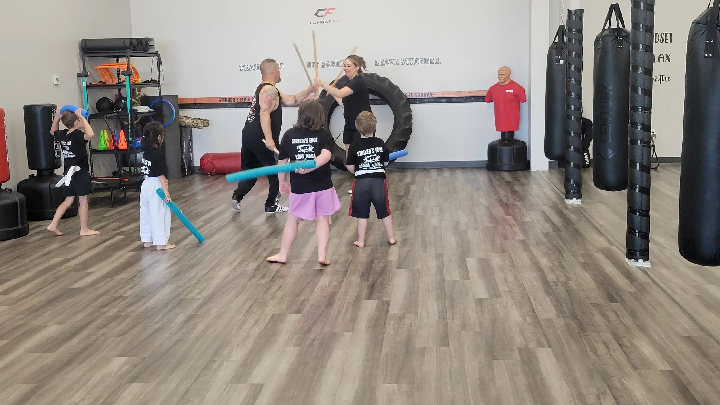Children with foam noodles watching two adults practicing a martial arts demonstration with sticks in a gym.