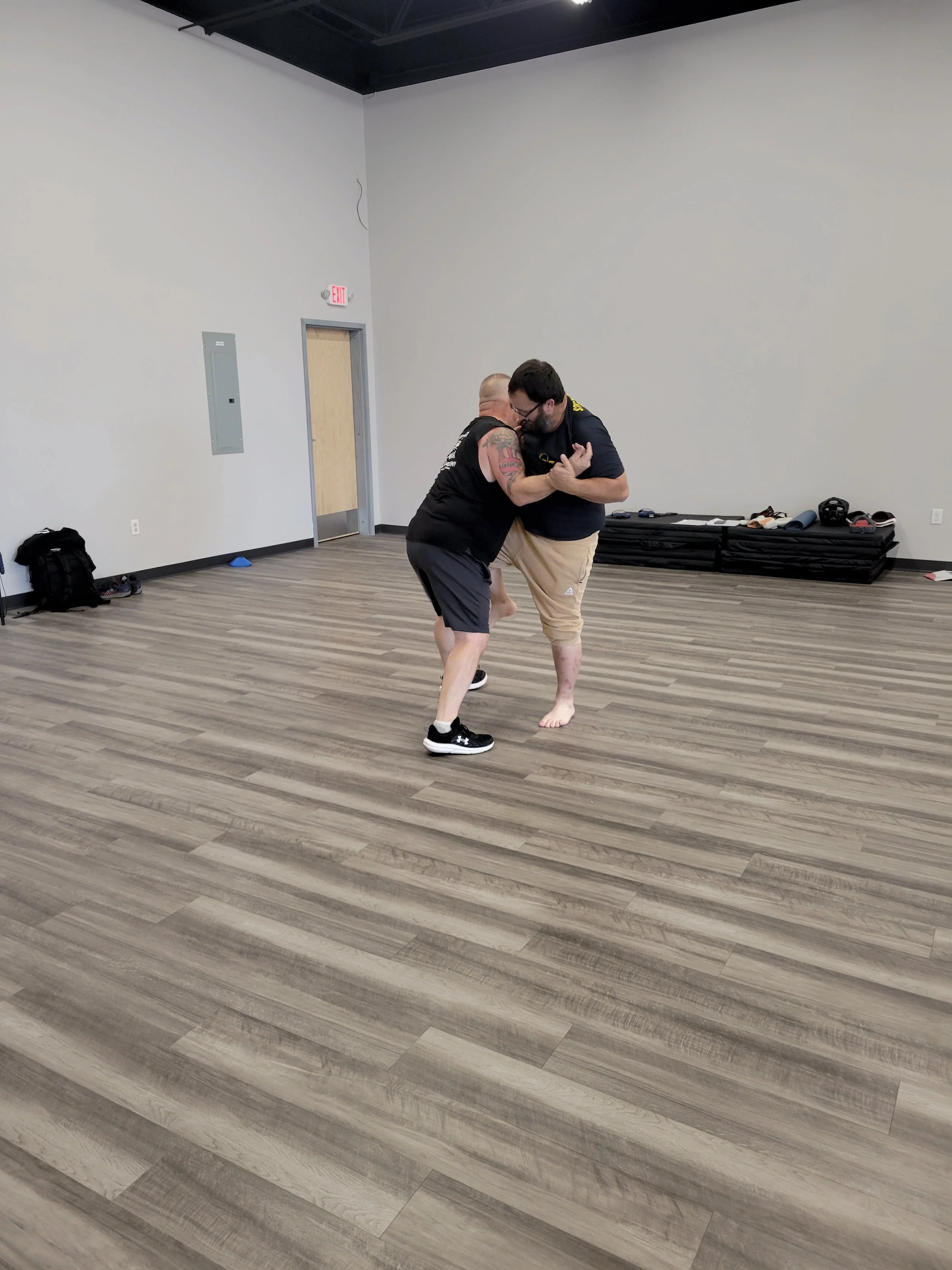 Two men practicing self-defense in an empty room with wood flooring, one man is holding the other's arm while they engage in close combat.