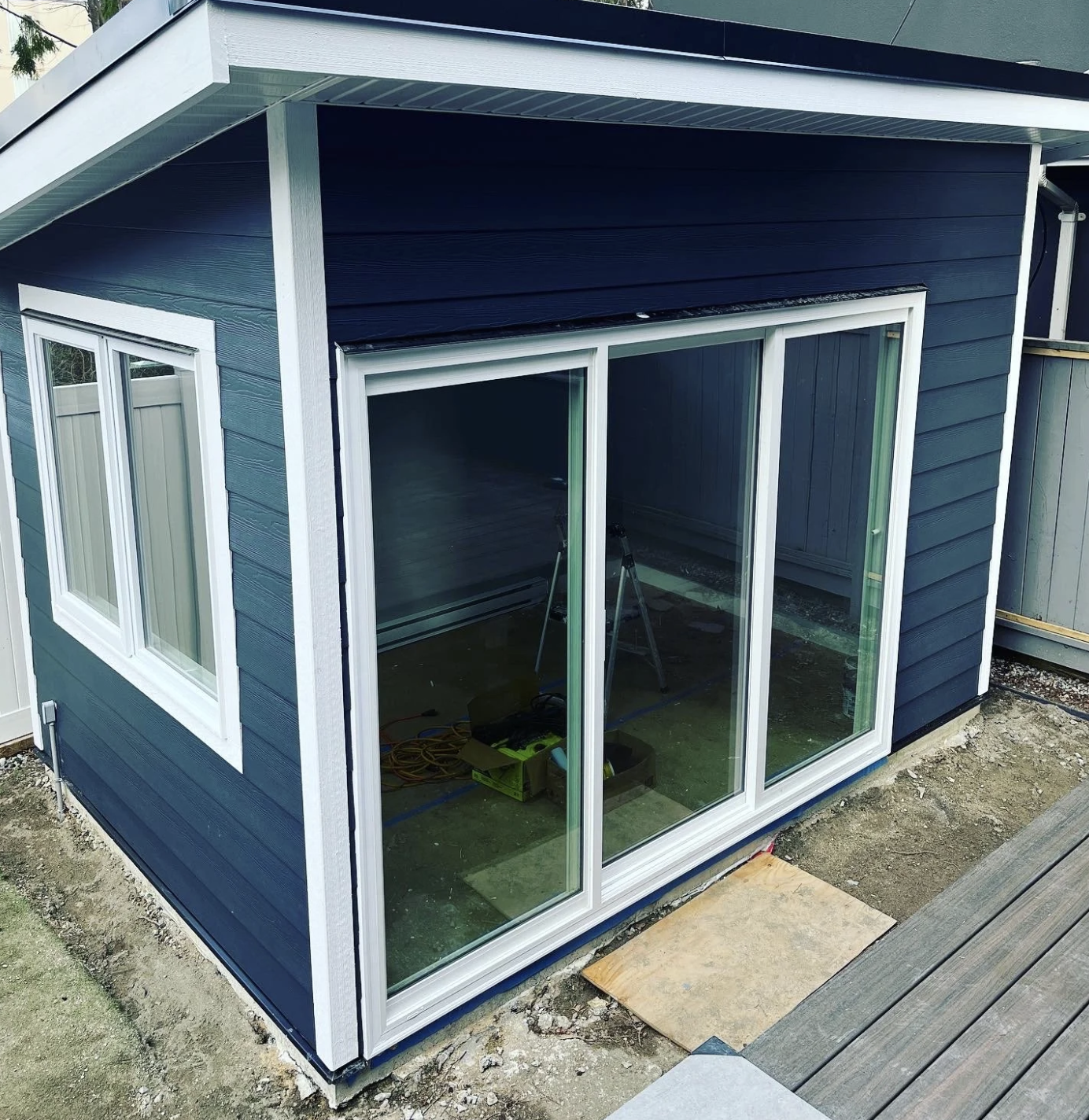 A small building under construction with navy blue siding, white-trimmed windows, and a sliding glass door. The ground around it is unfinished, with construction tools and materials inside.