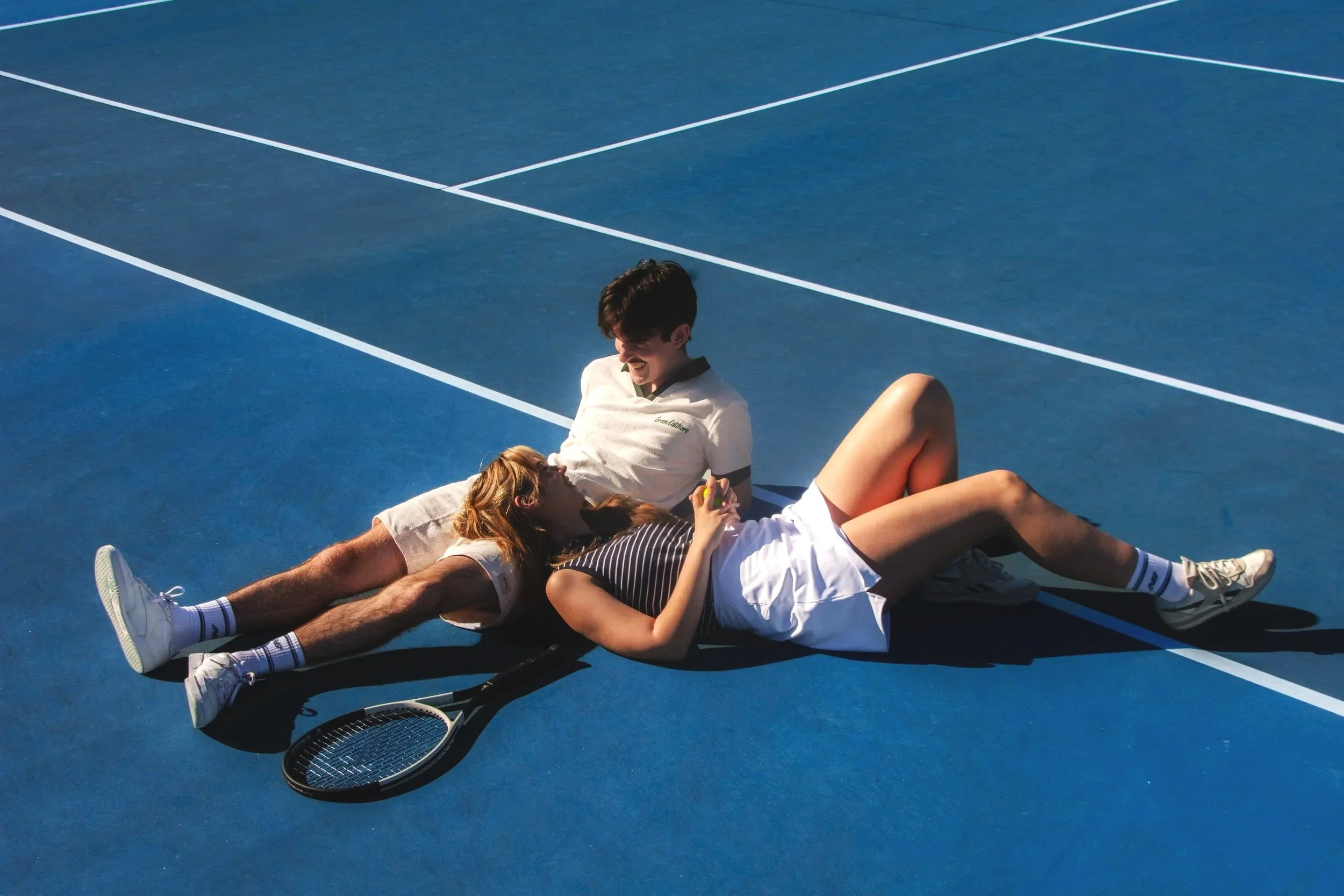 Two young people, a man and a woman, lying on a blue tennis court, smiling at each other. The woman holds a tennis ball, with tennis rackets nearby.