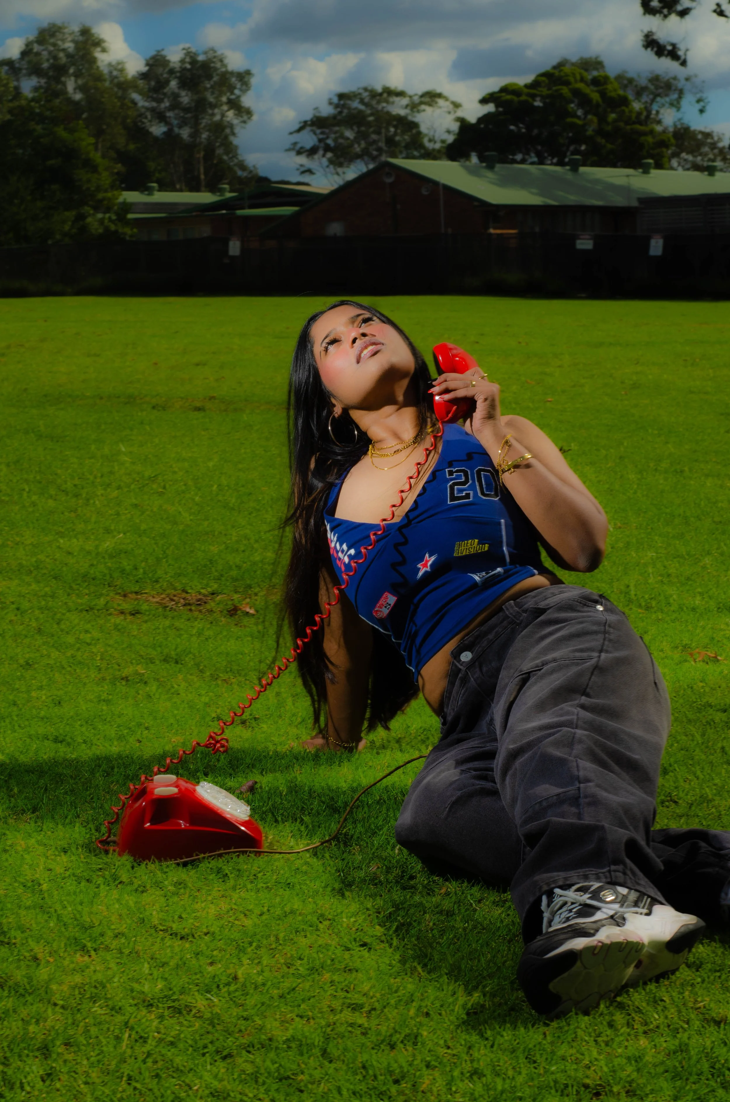 A woman is lying on the grass in a park, holding a red rotary telephone receiver to her ear. The cord of the telephone is attached to a red corded phone placed on the grass nearby. She is looking up with an expressive, slightly surprised or curious l