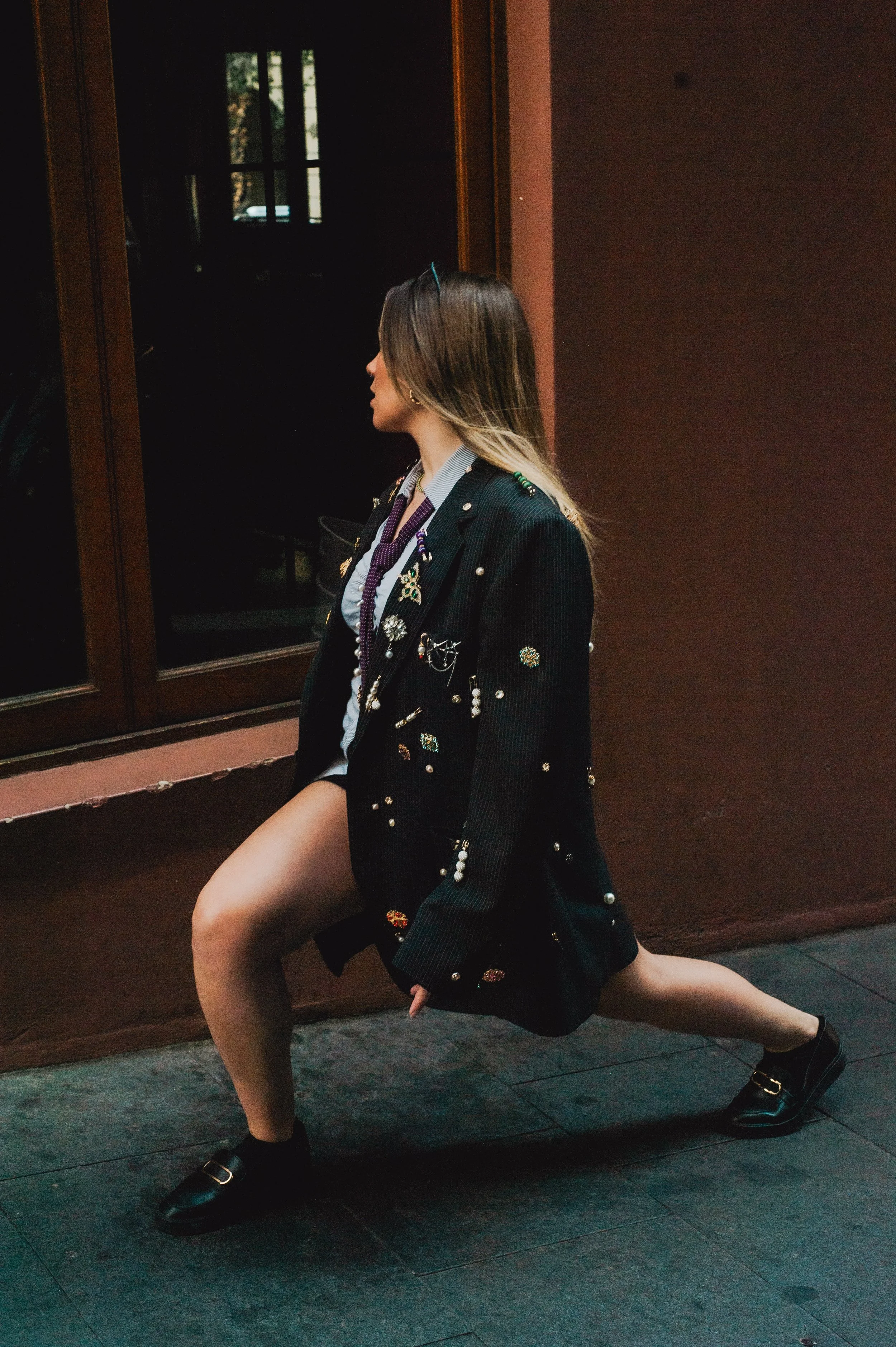 A woman in a black blazer decorated with various brooches, kneeling on one knee near a window with her hands on her knee, looking away from the camera.