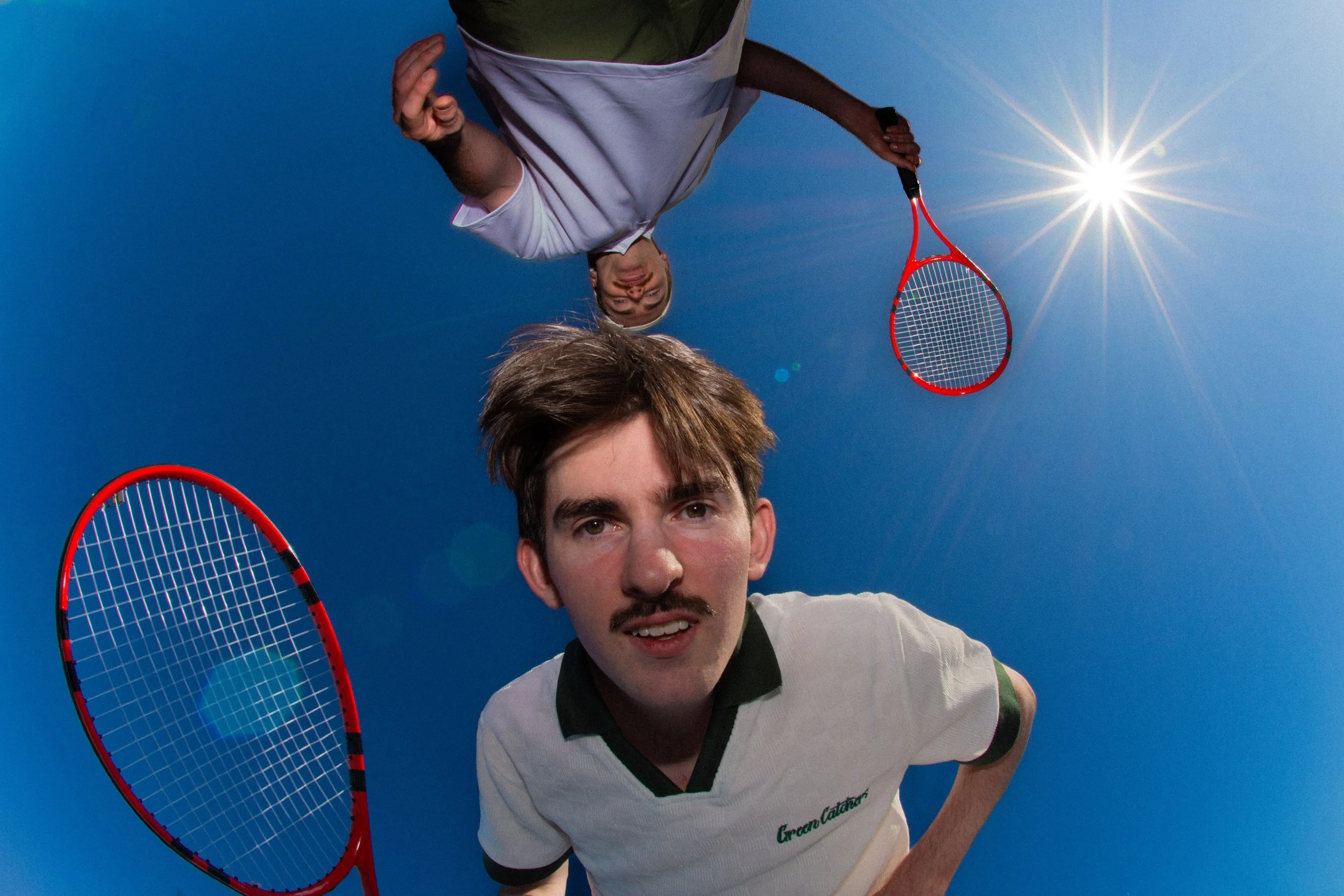 Two young men playing badminton outdoors on a sunny day with a clear blue sky, one holding a badminton racket and the other smiling at the camera.