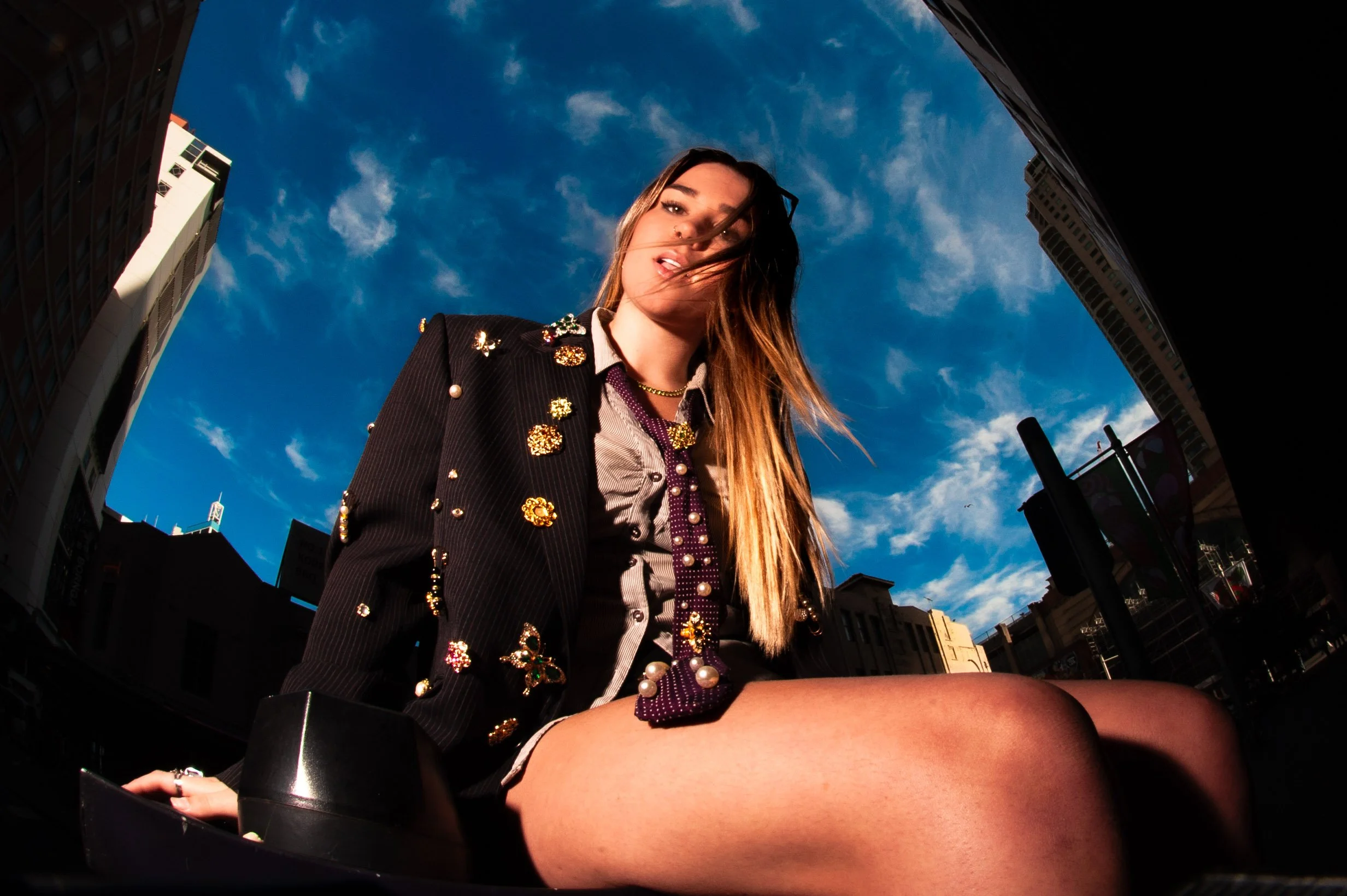 A woman sitting outdoors on a city street during daytime, wearing a decorated jacket, with a blue sky and tall buildings in the background.