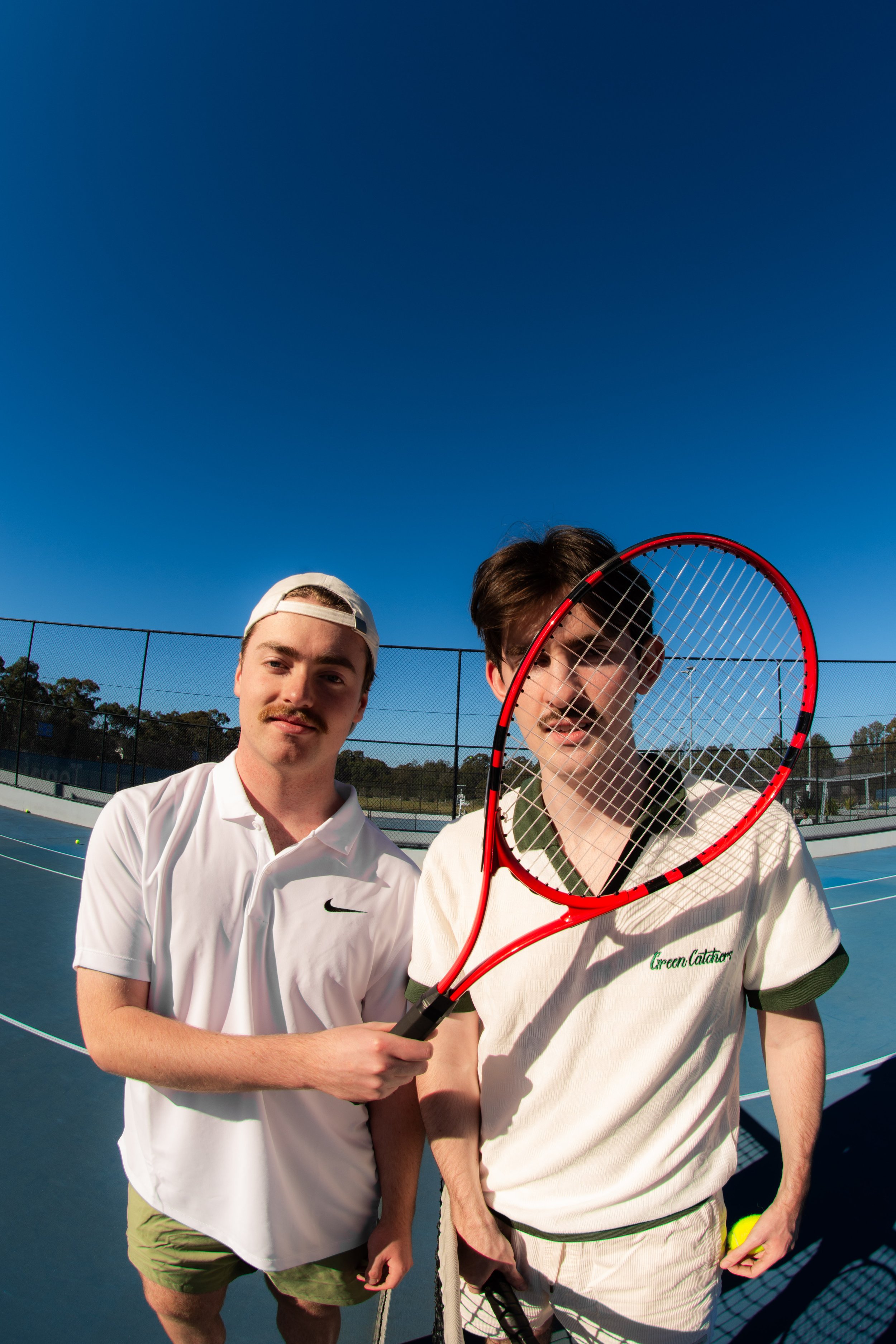 Two men on a tennis court, one holding a tennis racket in front of the other, with clear blue sky and a black fence in the background.