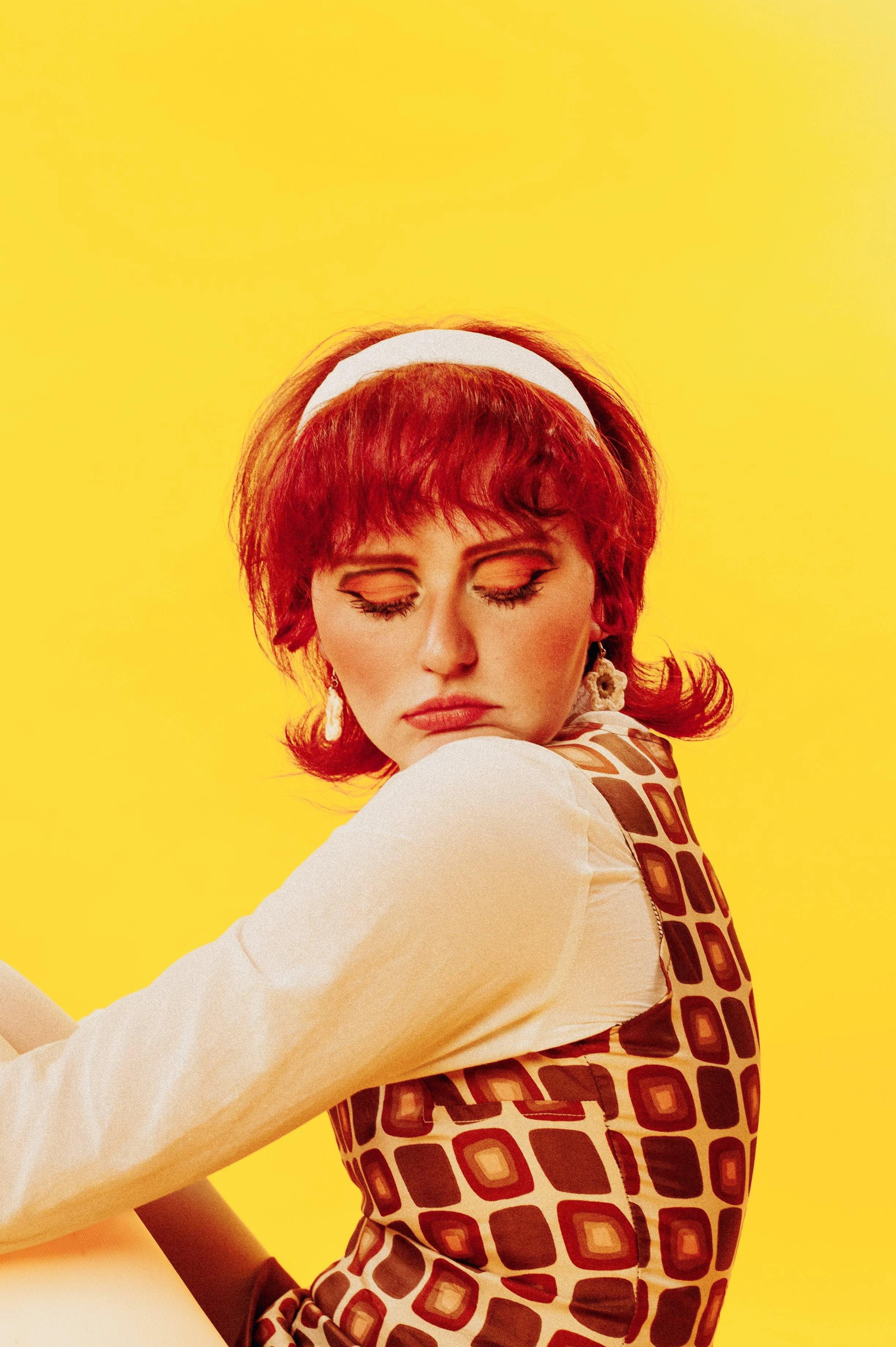 A woman with vibrant red hair, makeup, and earrings, wearing a white headband, a patterned top, and a long-sleeve shirt, with her eyes closed against a bright yellow background.