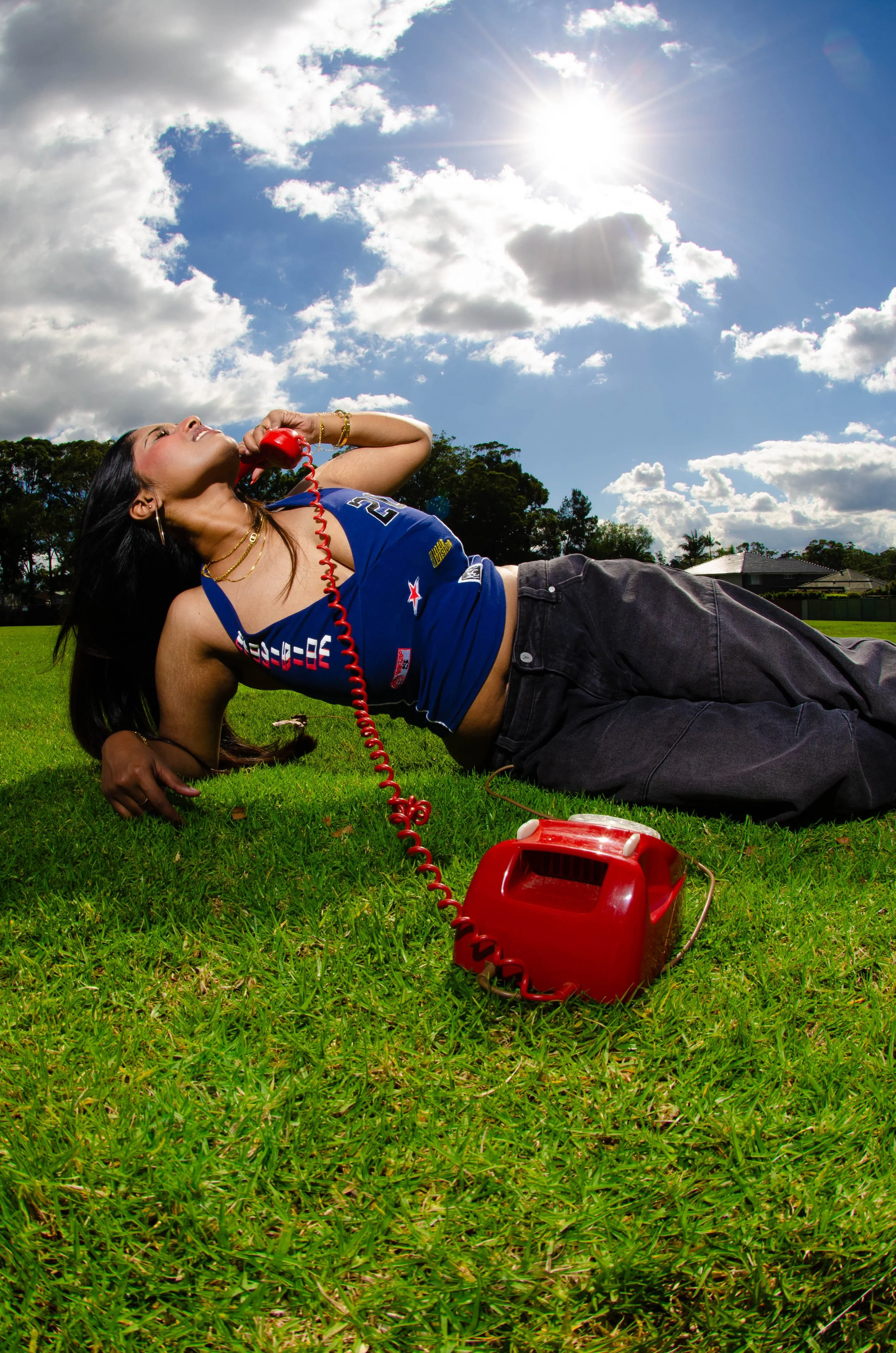 Woman lying on the grass outdoors talking on a red rotary phone, with a bright blue sky and sun overhead.