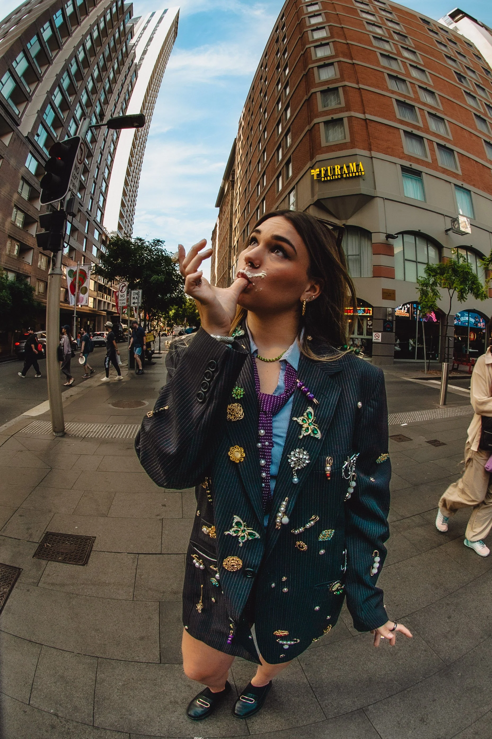A woman in a pinstriped suit jacket decorated with various pins and jewelry, standing on a city street, posing with her finger near her lips as she looks up. Tall buildings and pedestrians are visible in the background during daytime.