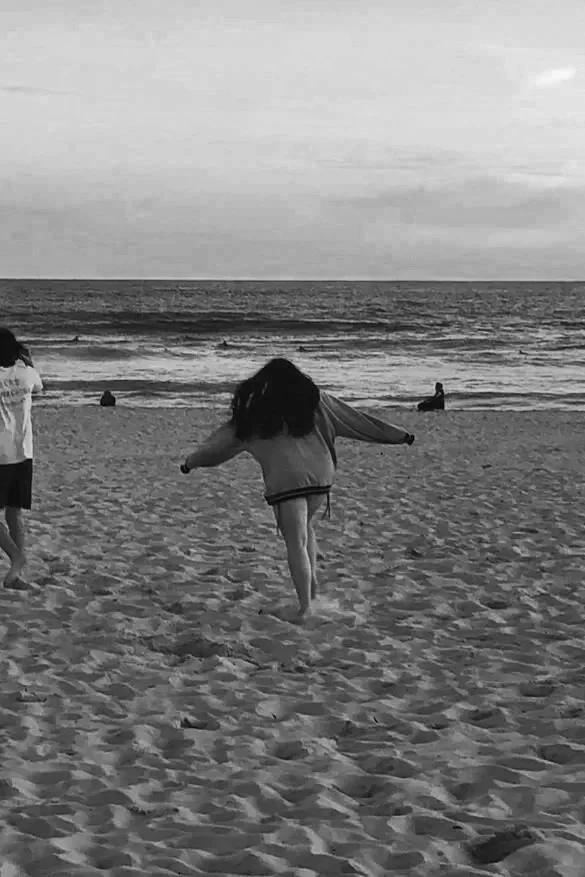 A black and white photo of a beach scene with a girl dancing or playing in the sand near the shoreline, with a few people sitting or standing in the water in the background.