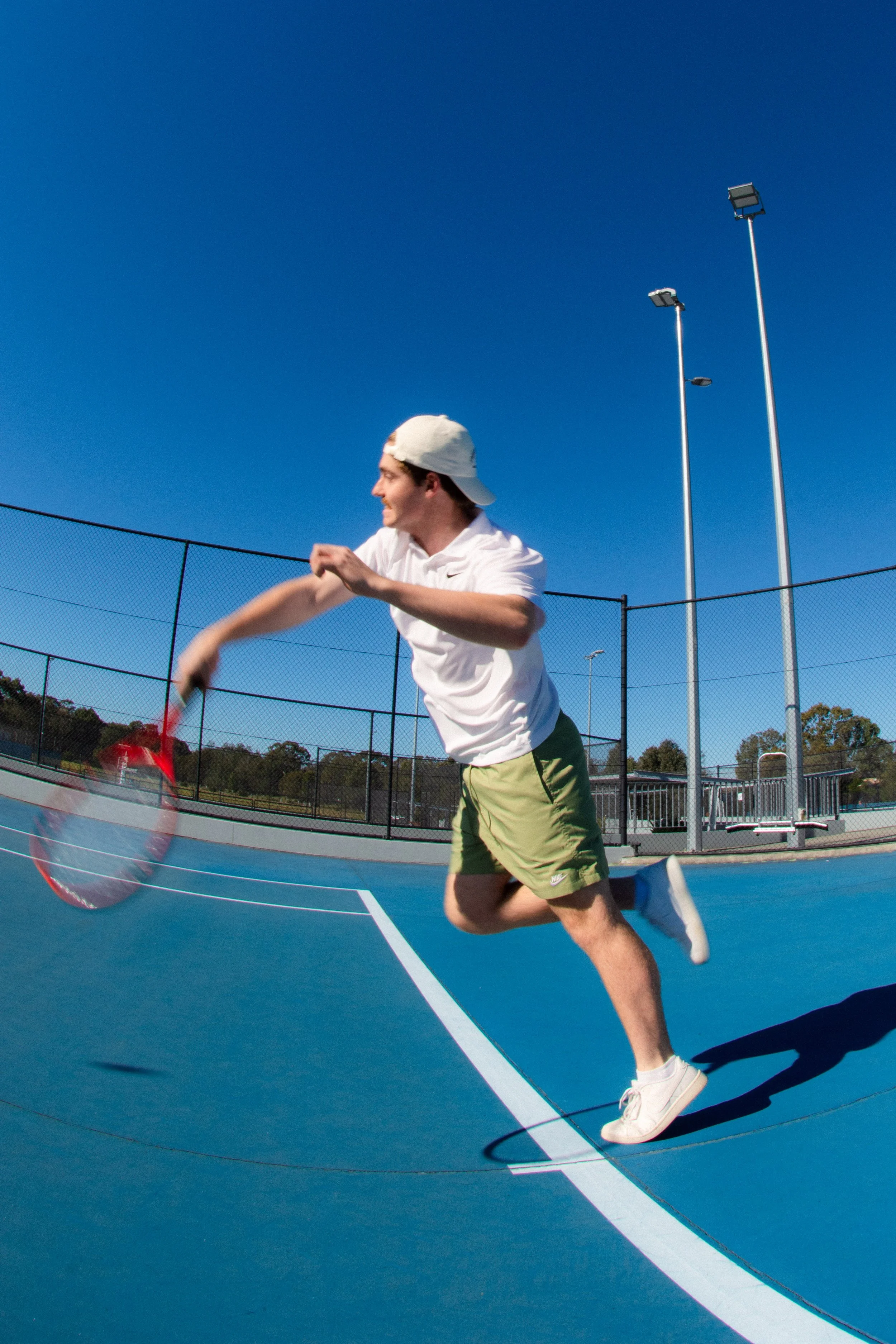 Young man in a white t-shirt, beige shorts, and a white cap playing pickleball on an outdoor court under a clear blue sky.