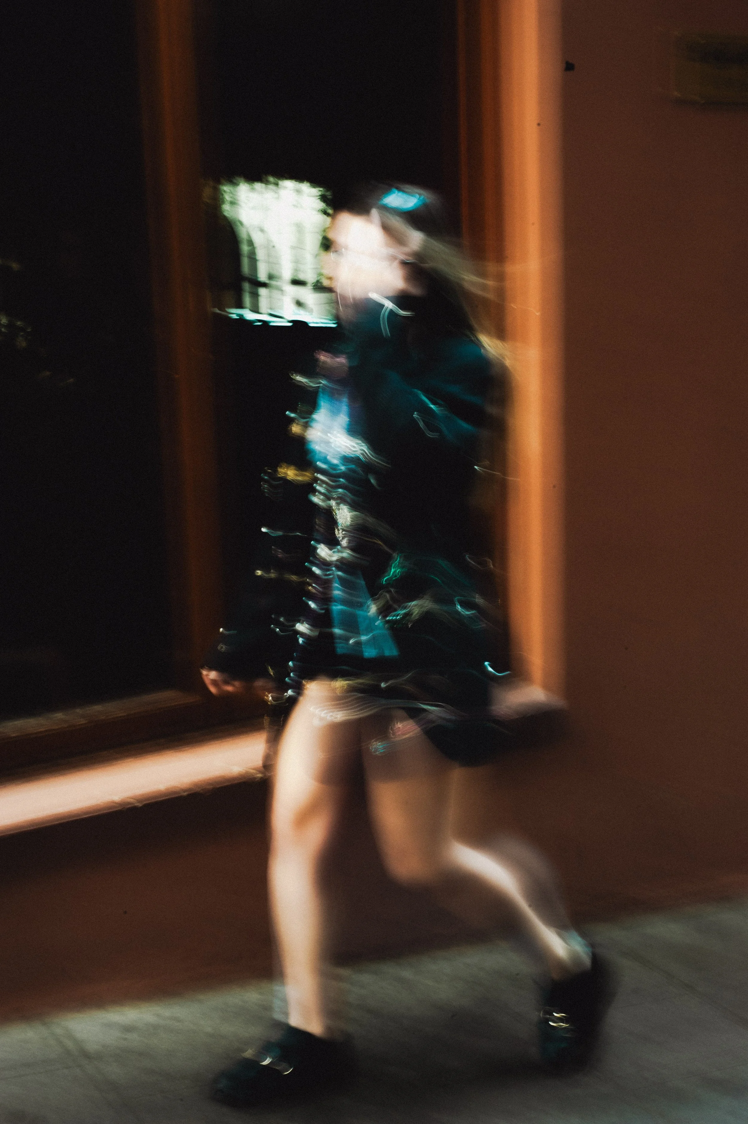 Blurred photo of a woman walking past a window at night, with visible light streaks and motion blur.