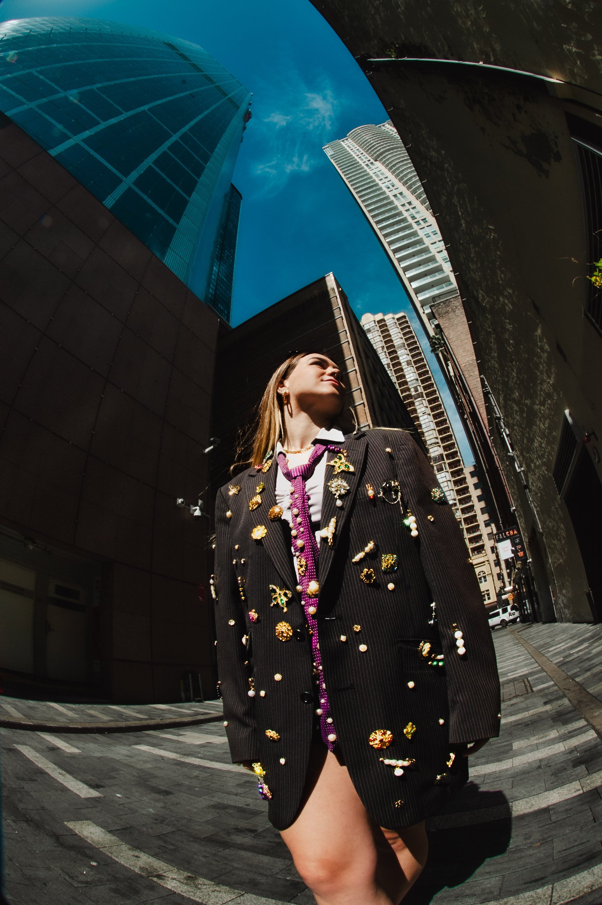 A woman in a black blazer jacket decorated with colorful jewelry stands on a city sidewalk with tall modern skyscrapers behind her, captured from a low-angle shot with fisheye lens under a clear blue sky.