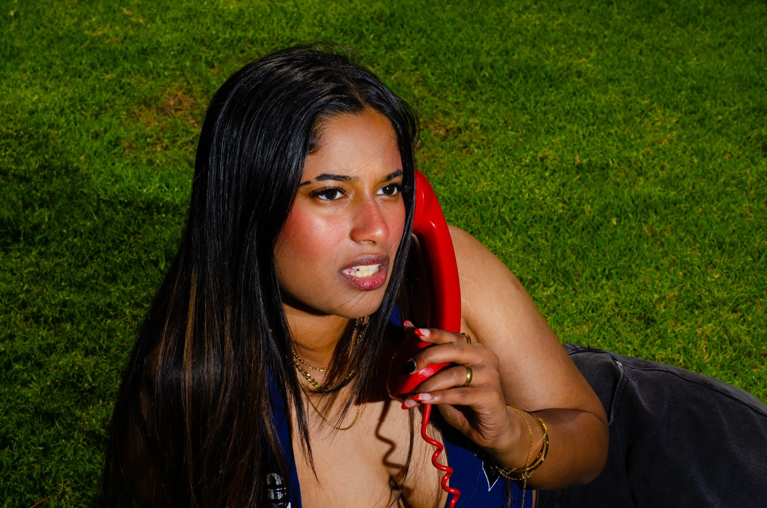 A woman with long dark hair lying on grass, holding a red telephone receiver to her ear, looking concerned.