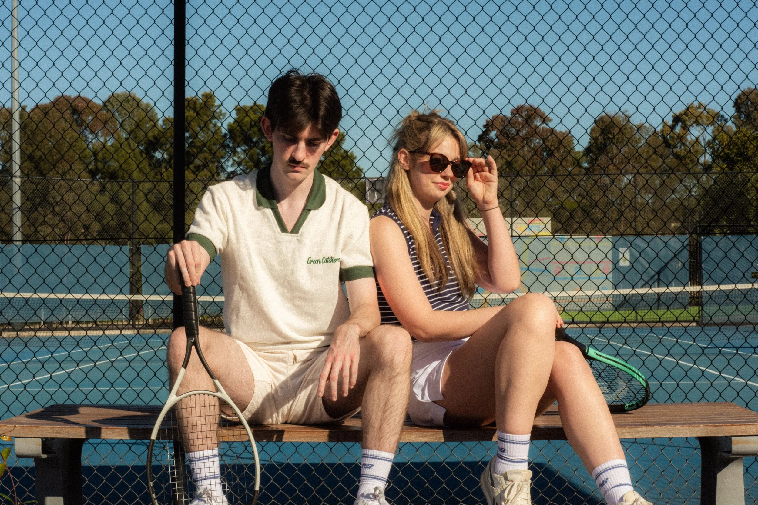 A young man and woman sitting on a bench by a tennis court, separated by a chain-link fence, with the man holding a tennis racket and the woman adjusting her sunglasses, both dressed in athletic wear on a sunny day.