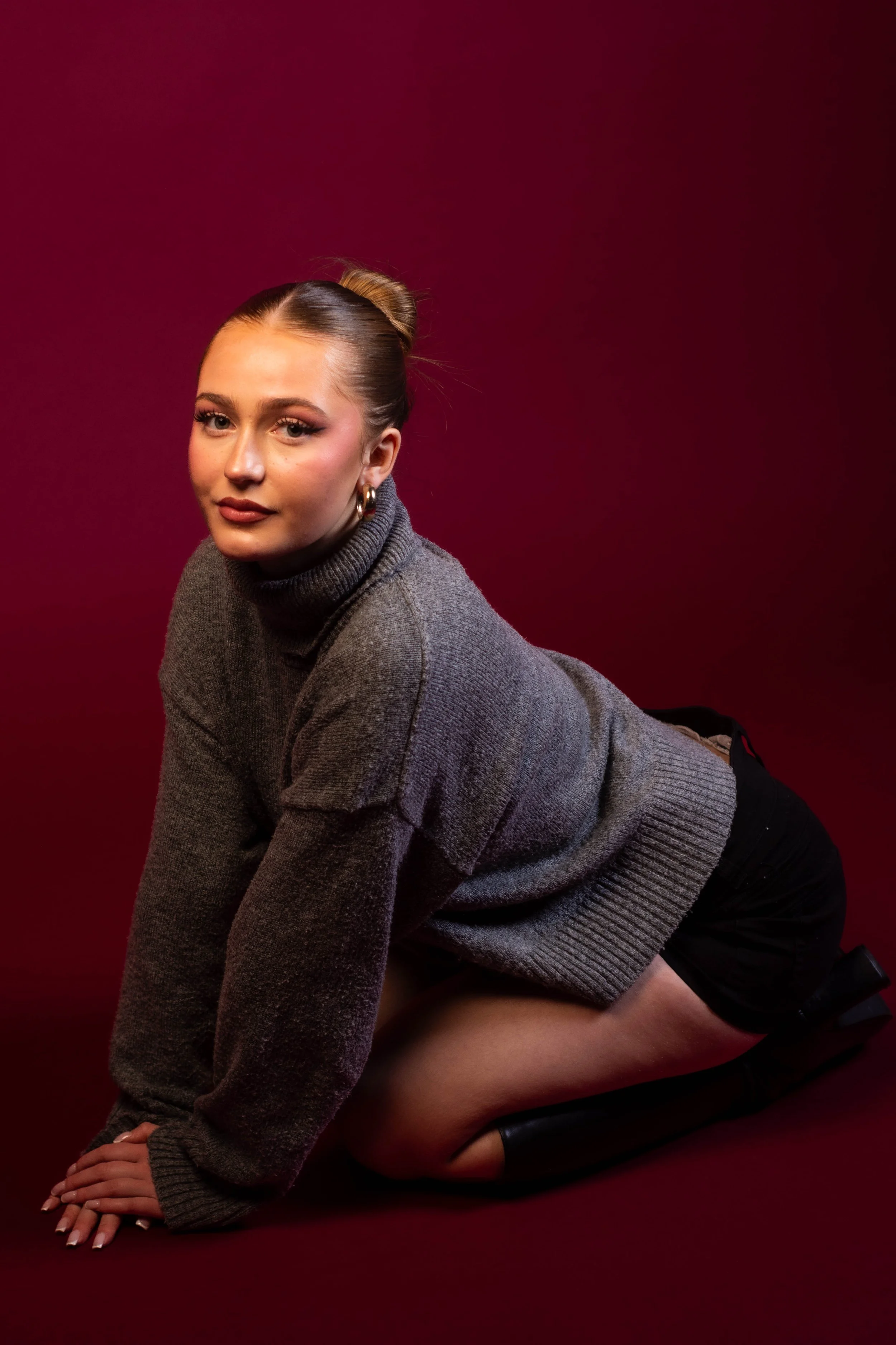 A woman kneeling on a red background, wearing a gray turtleneck sweater, black shorts, and thigh-high black boots, looking at the camera.