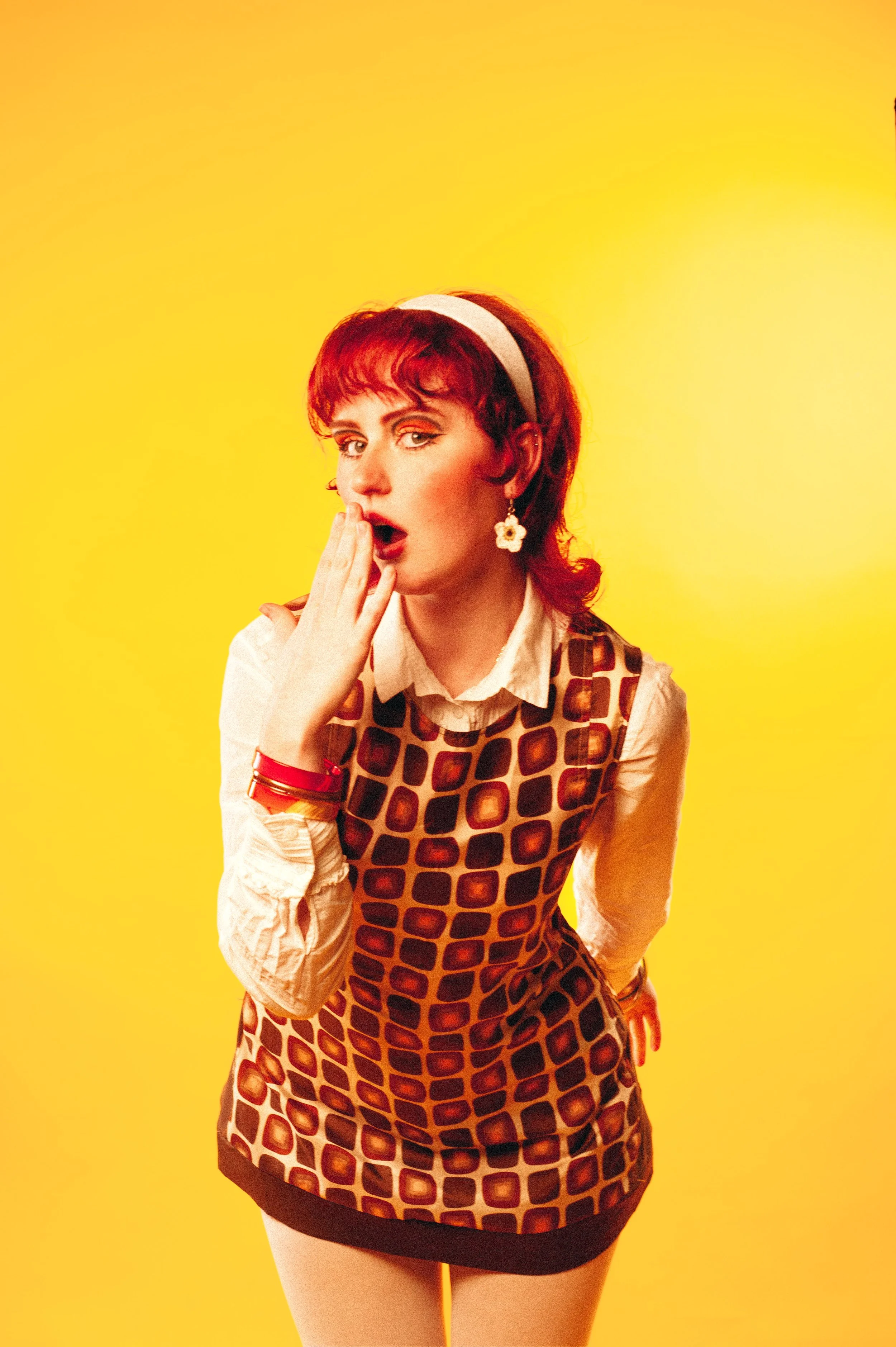 A woman with red hair and a headband, wearing a patterned dress over a white shirt, standing against a yellow background, with her mouth open and hand covering her mouth in surprise.