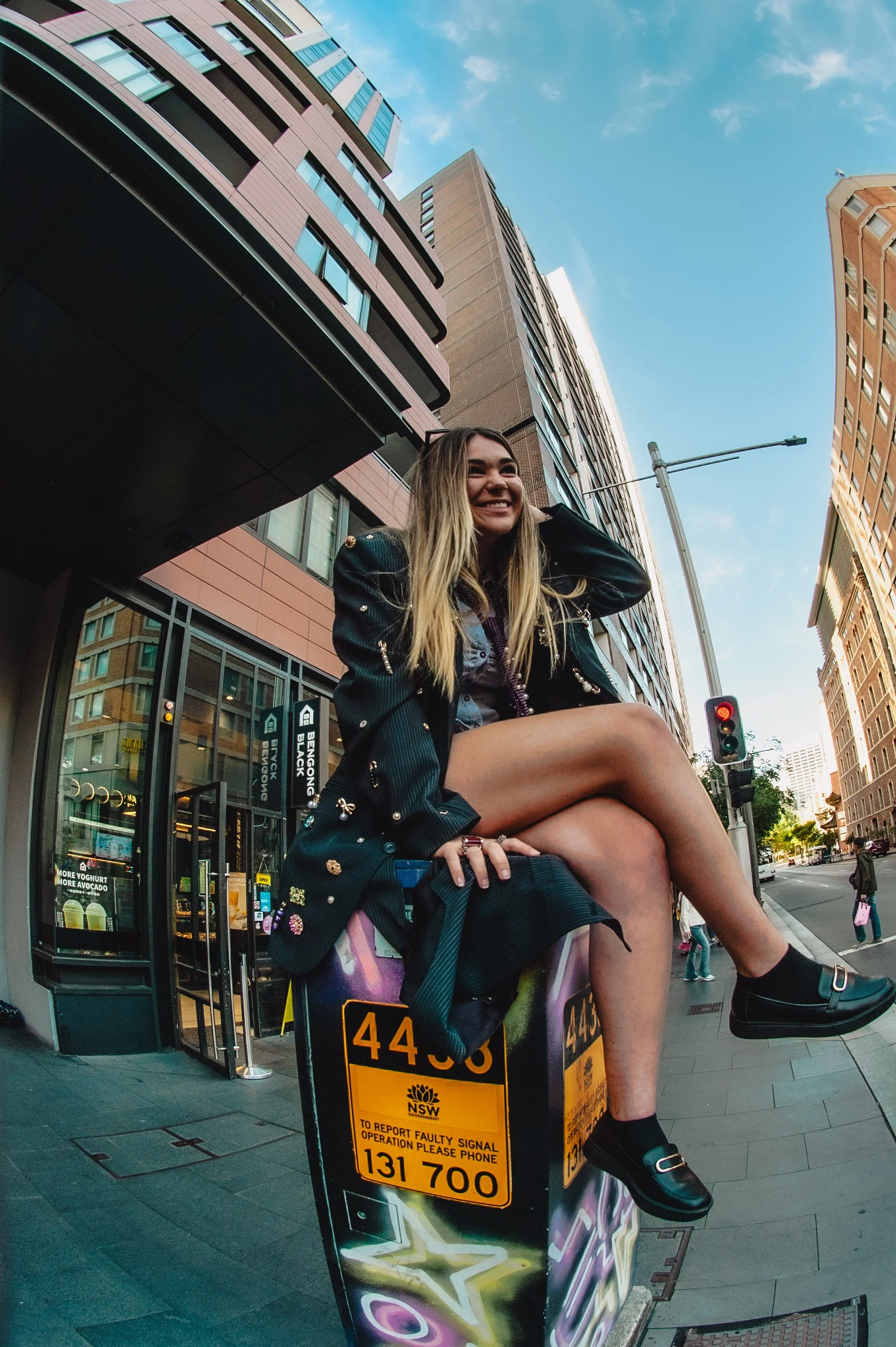 A young woman with long blonde hair sitting on a utility box on a city street corner, smiling, with tall buildings and a traffic light in the background.