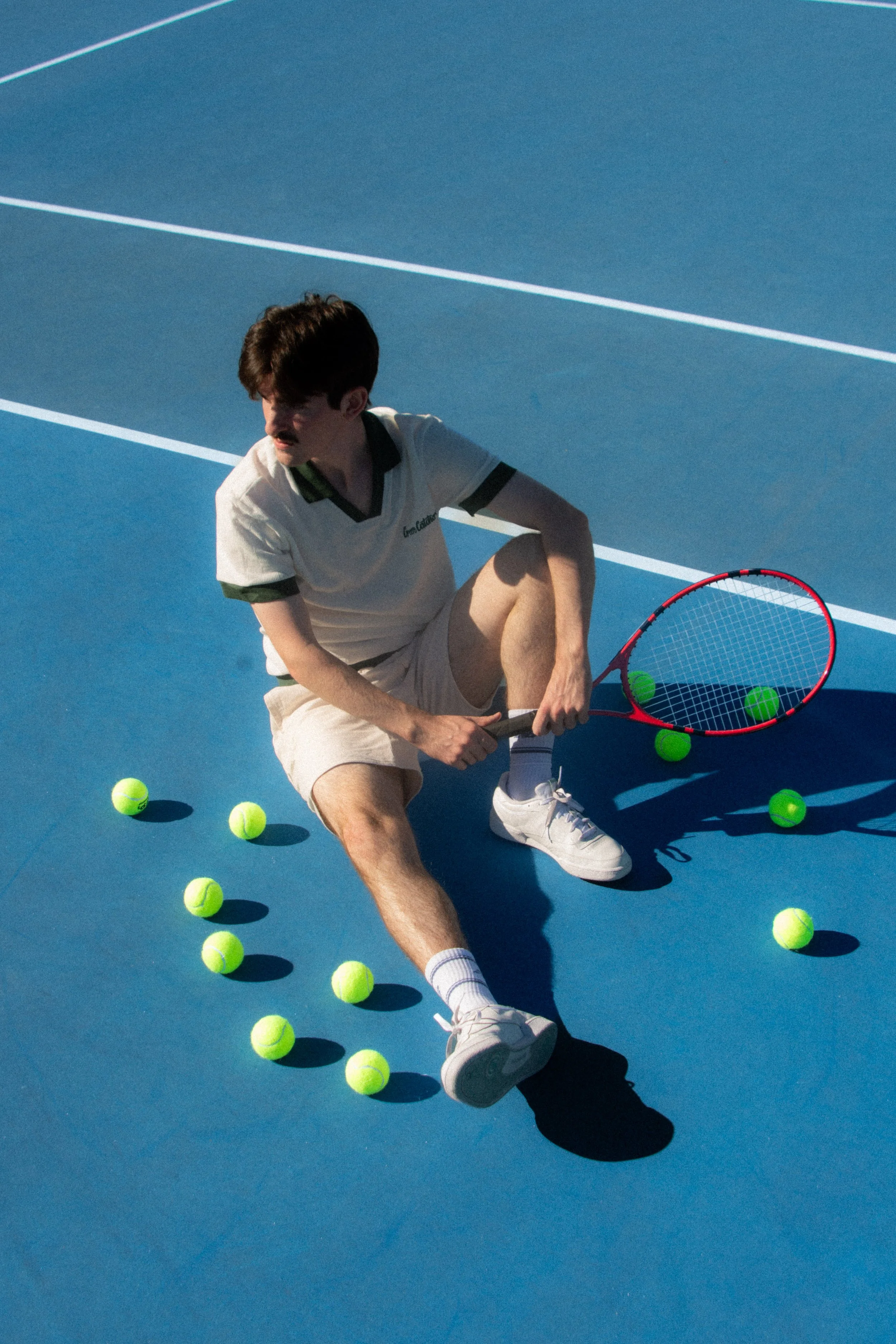 A young man sitting on a blue tennis court surrounded by tennis balls, with a tennis racket and three tennis balls inside the racket.