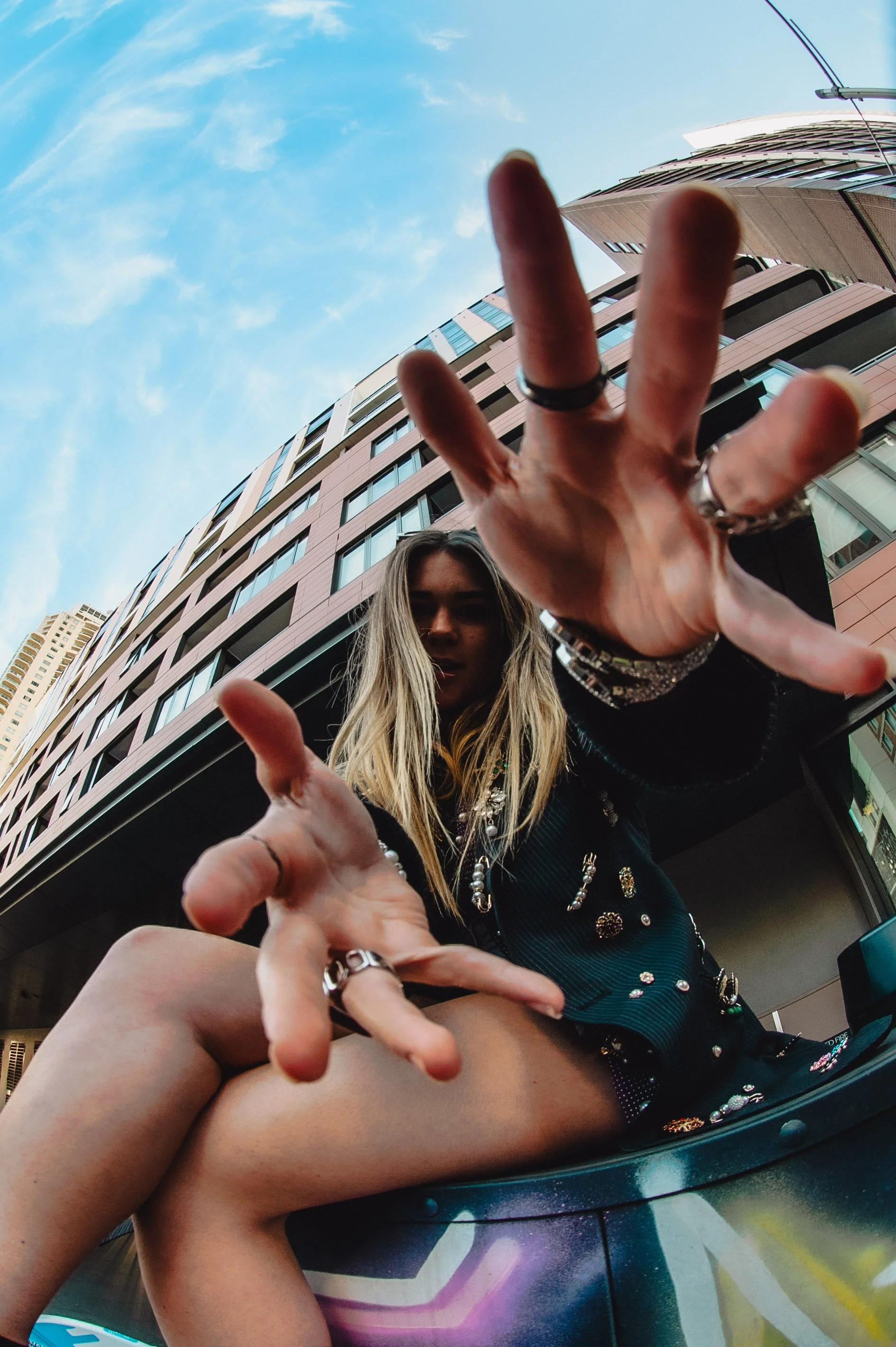 A woman with long blonde hair reaching toward the camera with two hands, taken from a low angle against a cityscape with tall buildings and a blue sky.