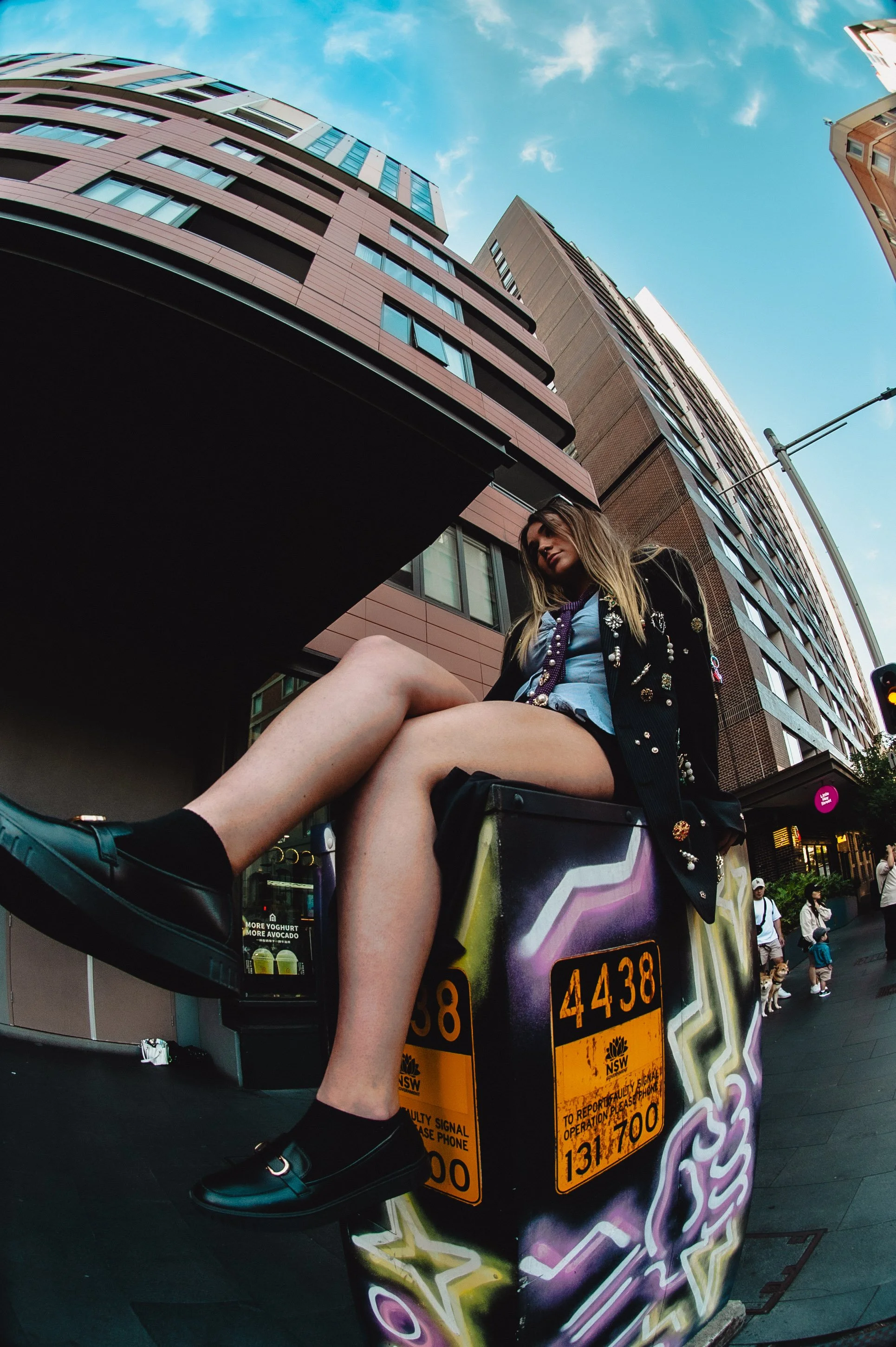 A young woman with long blonde hair sitting on a graffitied utility box on a city sidewalk, with tall modern buildings and a bright blue sky in the background.