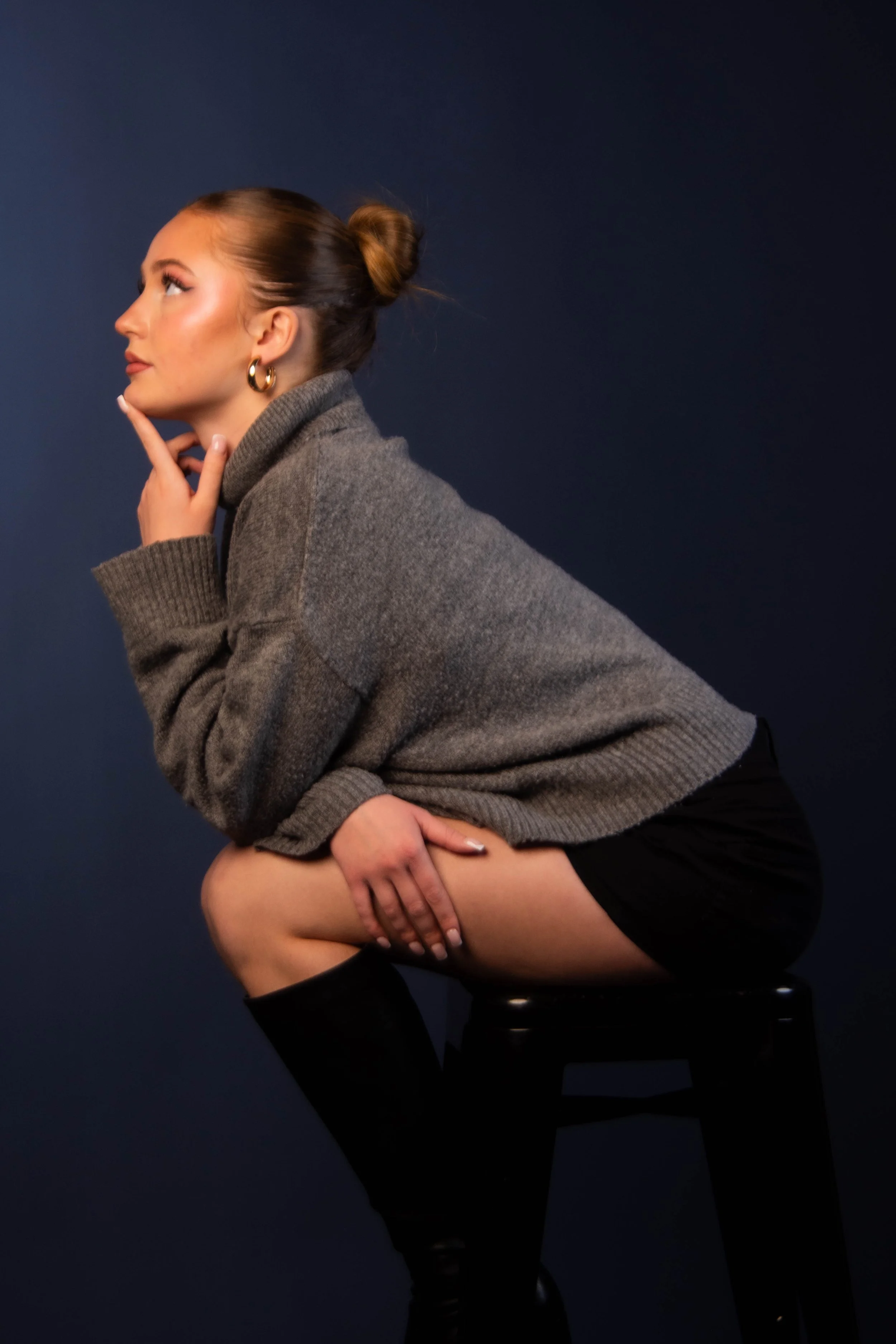 A young woman with makeup and hoop earrings, sitting on a stool, wearing a gray sweater and black shorts, with her hair in a bun against a dark background.