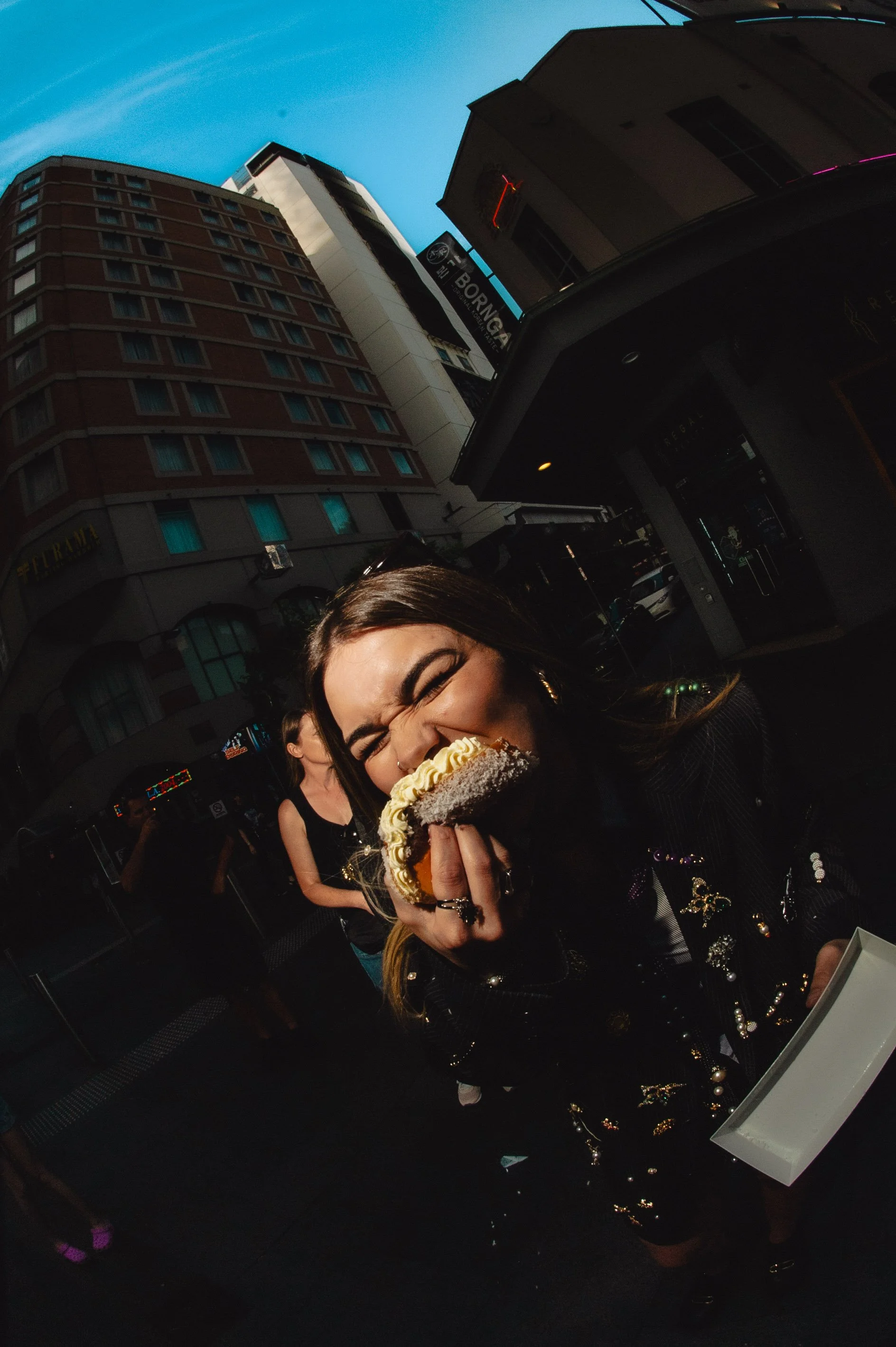 Young woman with long hair biting into a large slice of cake outdoors in a city setting, with buildings and pedestrians in the background.
