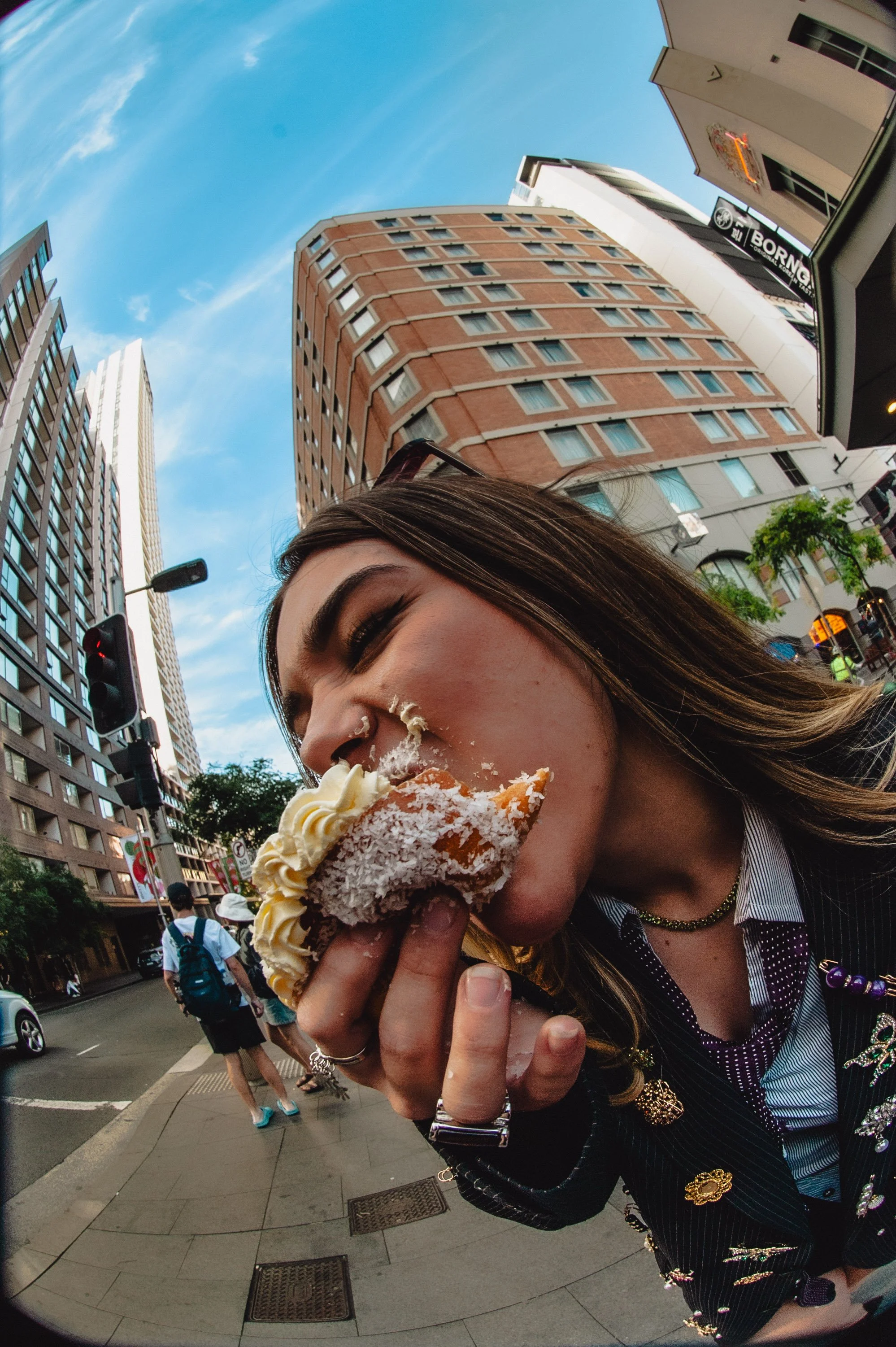 Young woman with long brown hair eating a slice of coconut cake topped with whipped cream on a city street with tall buildings and a blue sky.