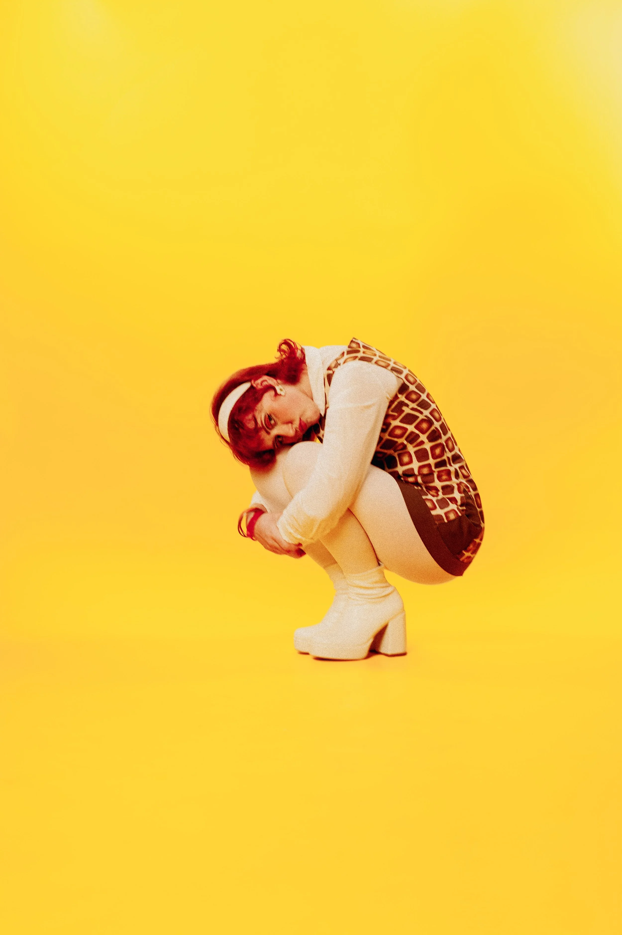A woman with red hair and a white headband, dressed in retro-style clothing, is squatting on a bright yellow background, hugging her knees and looking down.