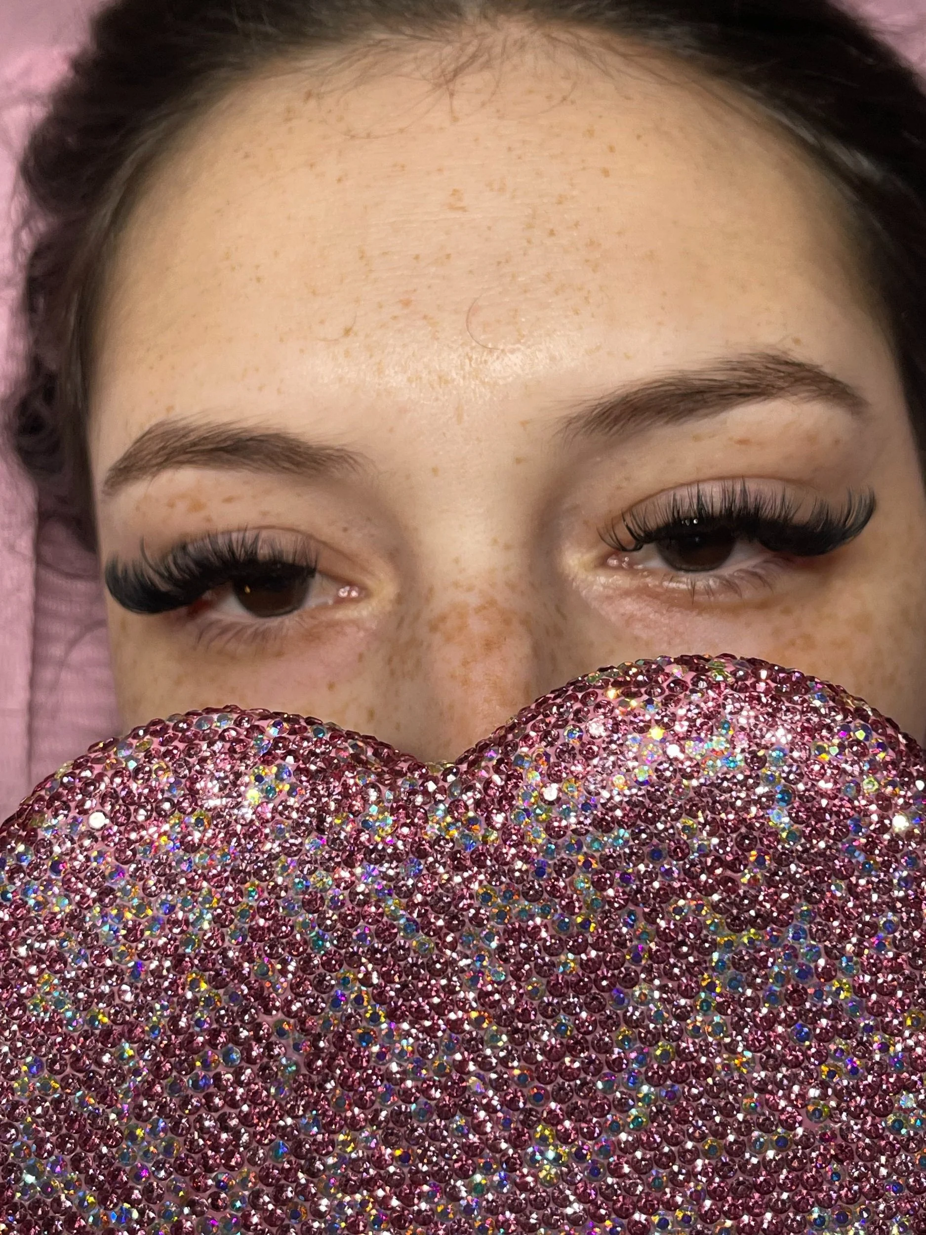 A close-up of a woman with valkyrie-style false eyelashes, freckles on her face, and dark hair, holding a heart-shaped object covered in pink and multicolored rhinestones in front of her face.