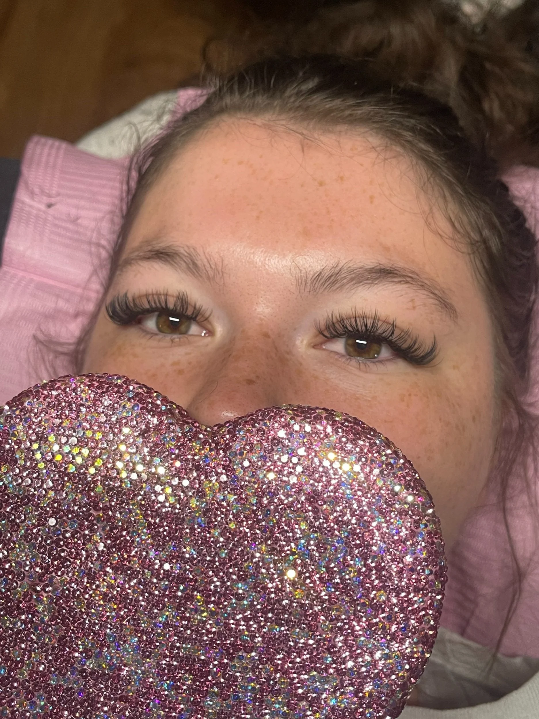 Close-up of a young woman with brown eyes, long eyelash extensions, freckles on her face, lying down with her head on a pink pillow, holding a large pink heart-shaped object covered in rhinestones in front of her face.