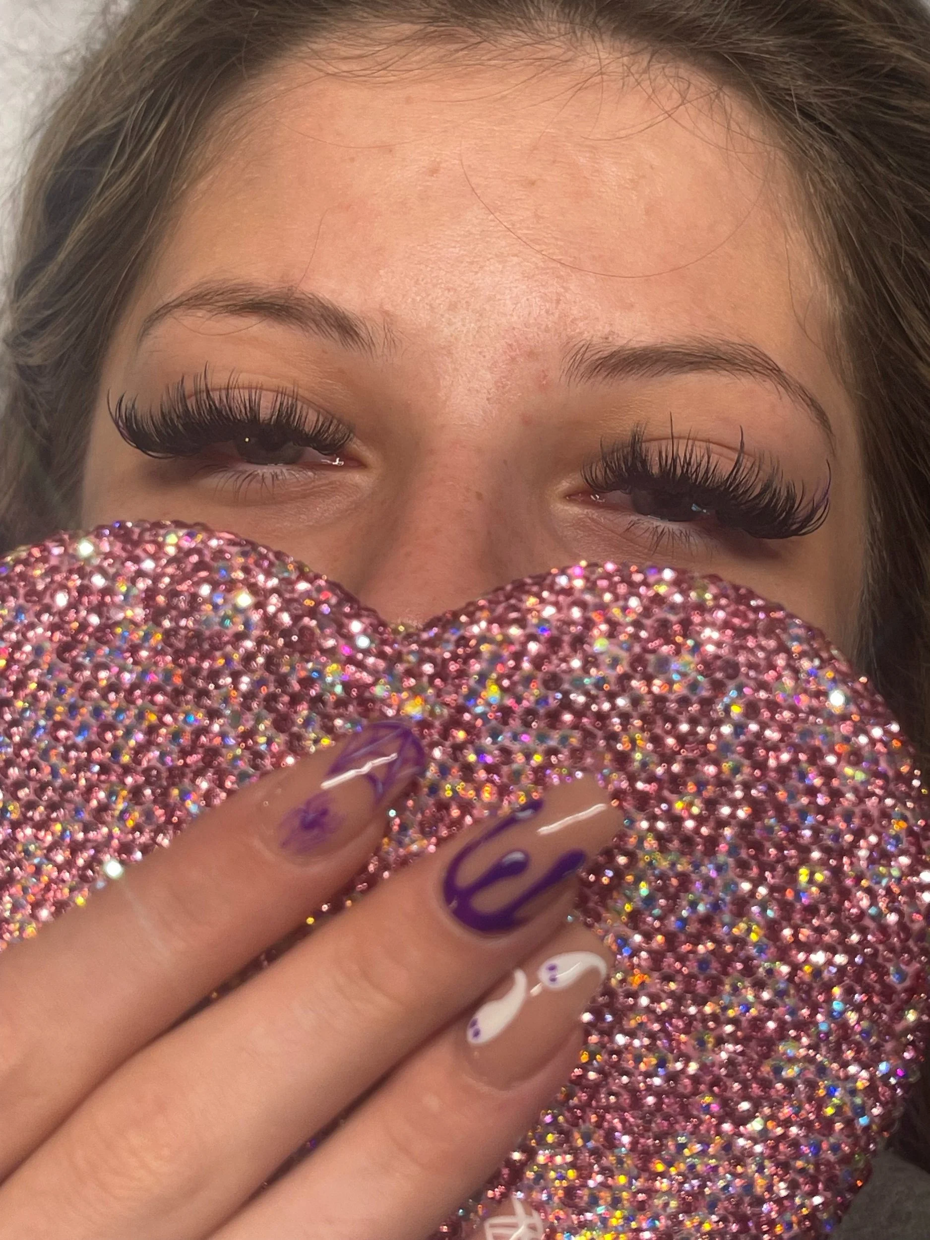 A woman with long false eyelashes holding a heart-shaped, glittery pink object close to her face, with her eyes partially closed.