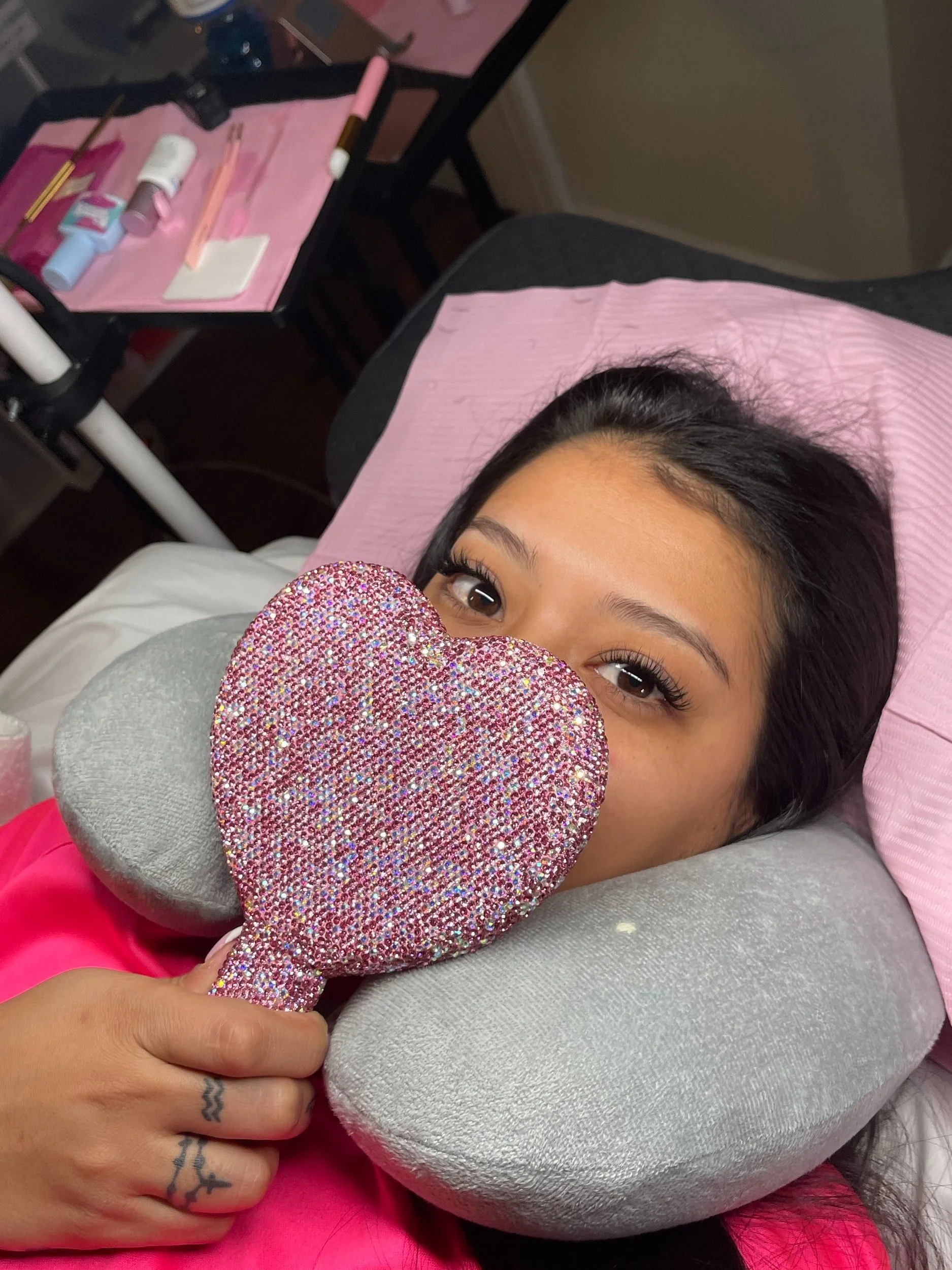 A woman lying on a bed with a pink pillow, holding a sparkly heart-shaped pillow in front of her face and resting her head on a neck pillow, with a table of nail supplies in the background.